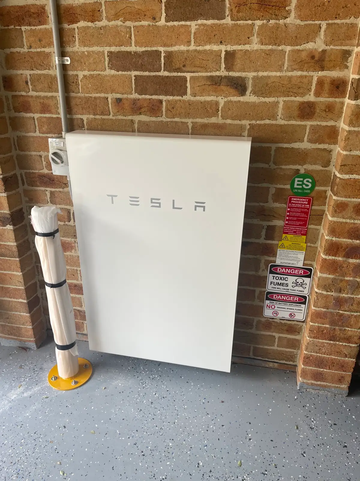A Tesla battery against a brick garage wall, hazard placards and an isolator beside it.