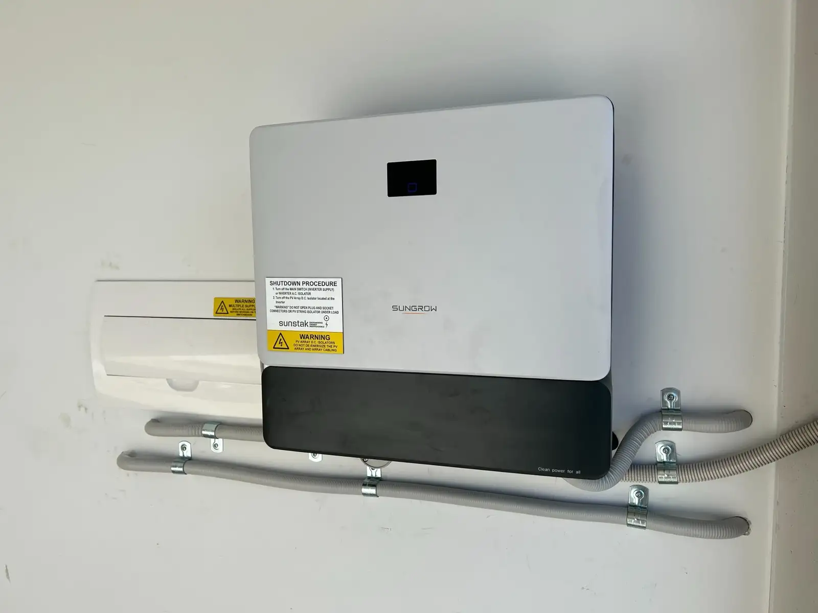 A Sungrow inverter on a white wall carrying a Sunstak shutdown label, conduit clipped in straight lines below.