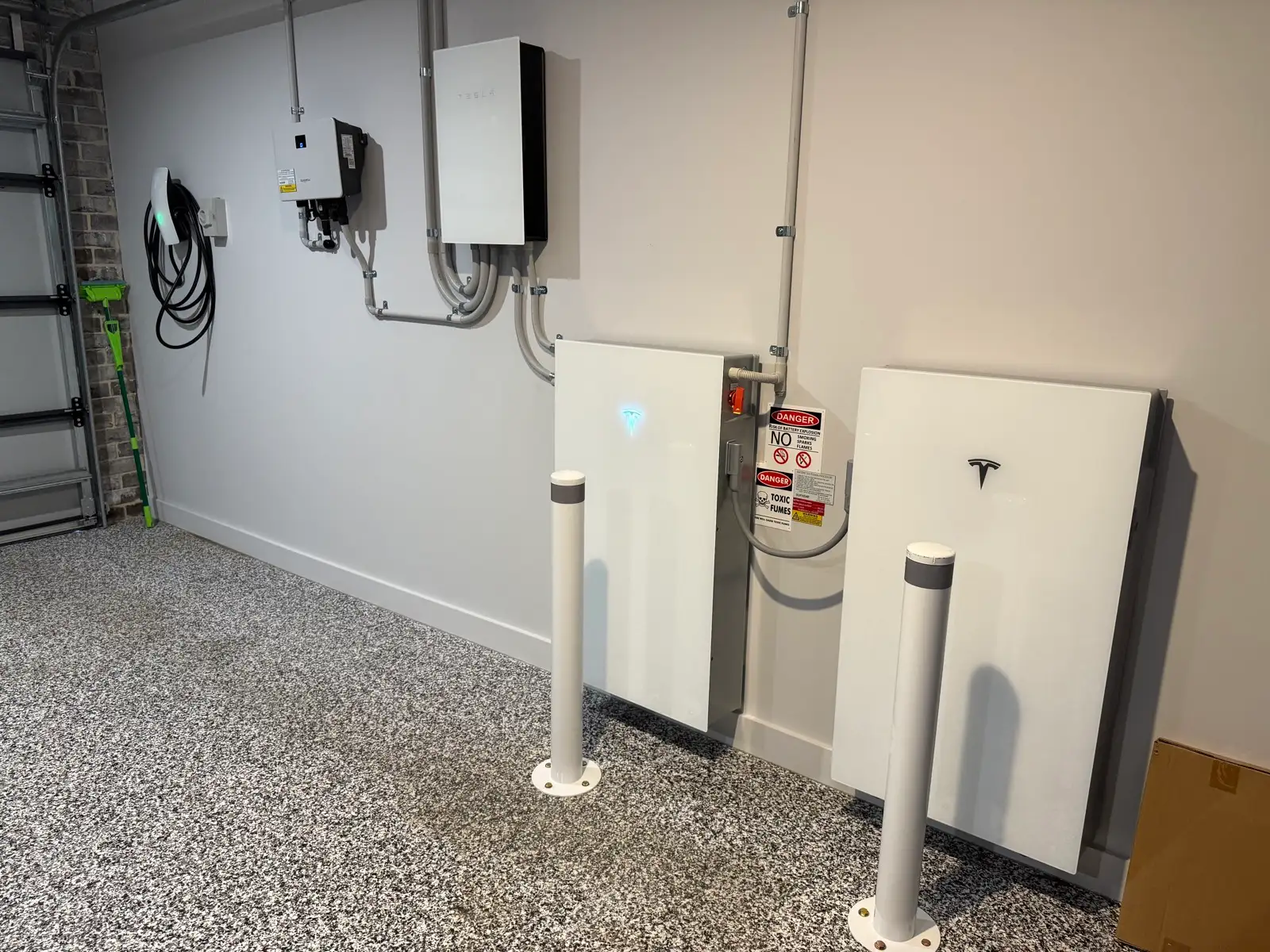 Two Tesla batteries on a garage floor behind bollards, the switchgear and an EV charger on the wall above.
