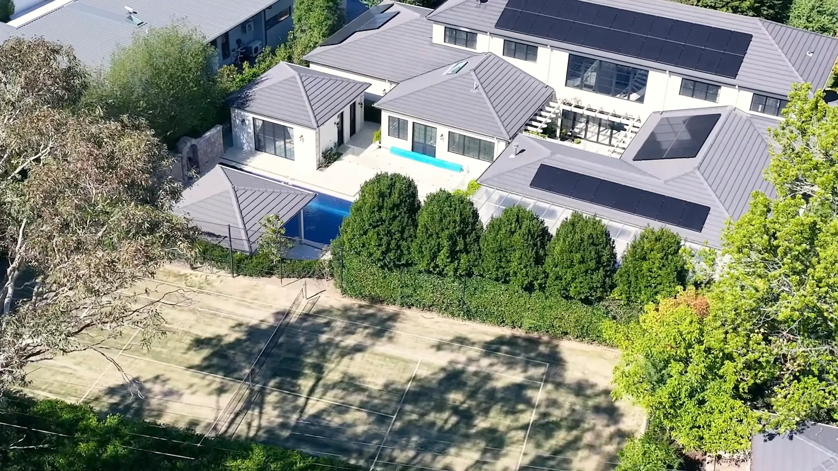 Dark solar panels laid across three separate faces of a grey metal roof, seen from the air, with a blue lap pool and a courtyard between the wings of the house below