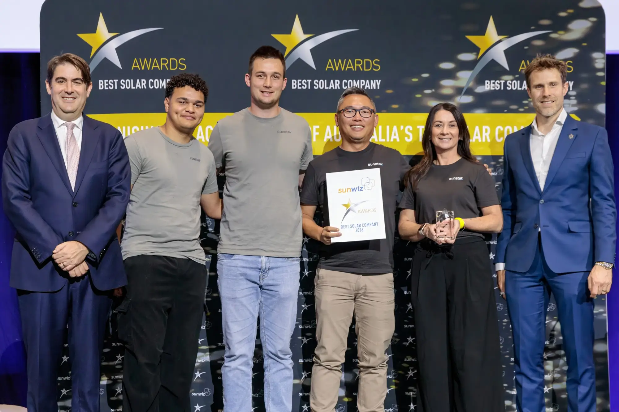 Six people in Sunstak T-shirts and suits on a stage in front of a navy ‘Best Solar Company’ awards backdrop, one holding a SunWiz Best Solar Company 2026 certificate and a glass trophy