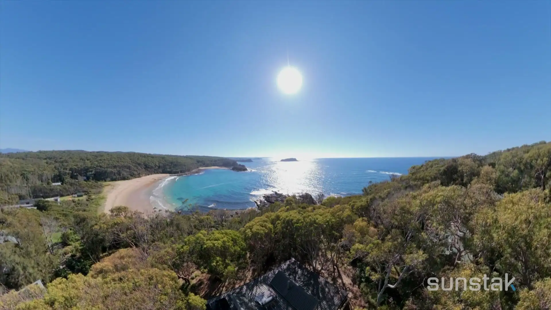 A wide view from above the treetops: a crescent of pale sand and turquoise water below a headland, a small rocky island offshore, open ocean to the horizon with the sun low over it, and the dark metal roof of a house in the bushland in the foreground