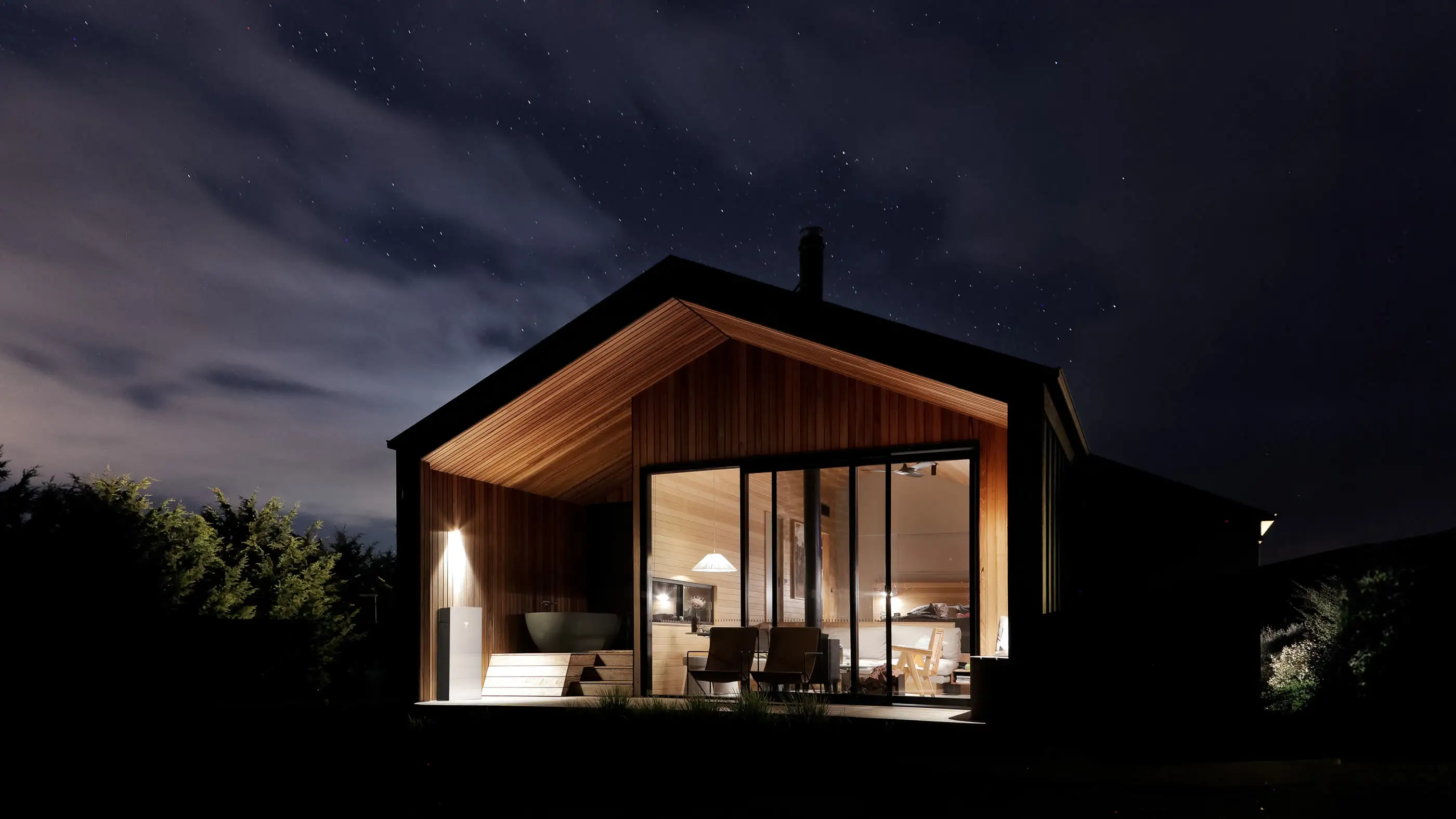 A black timber cabin with a steep gable at night under a starry sky, its glass end wall showing a warm lit room with armchairs and a bath inside