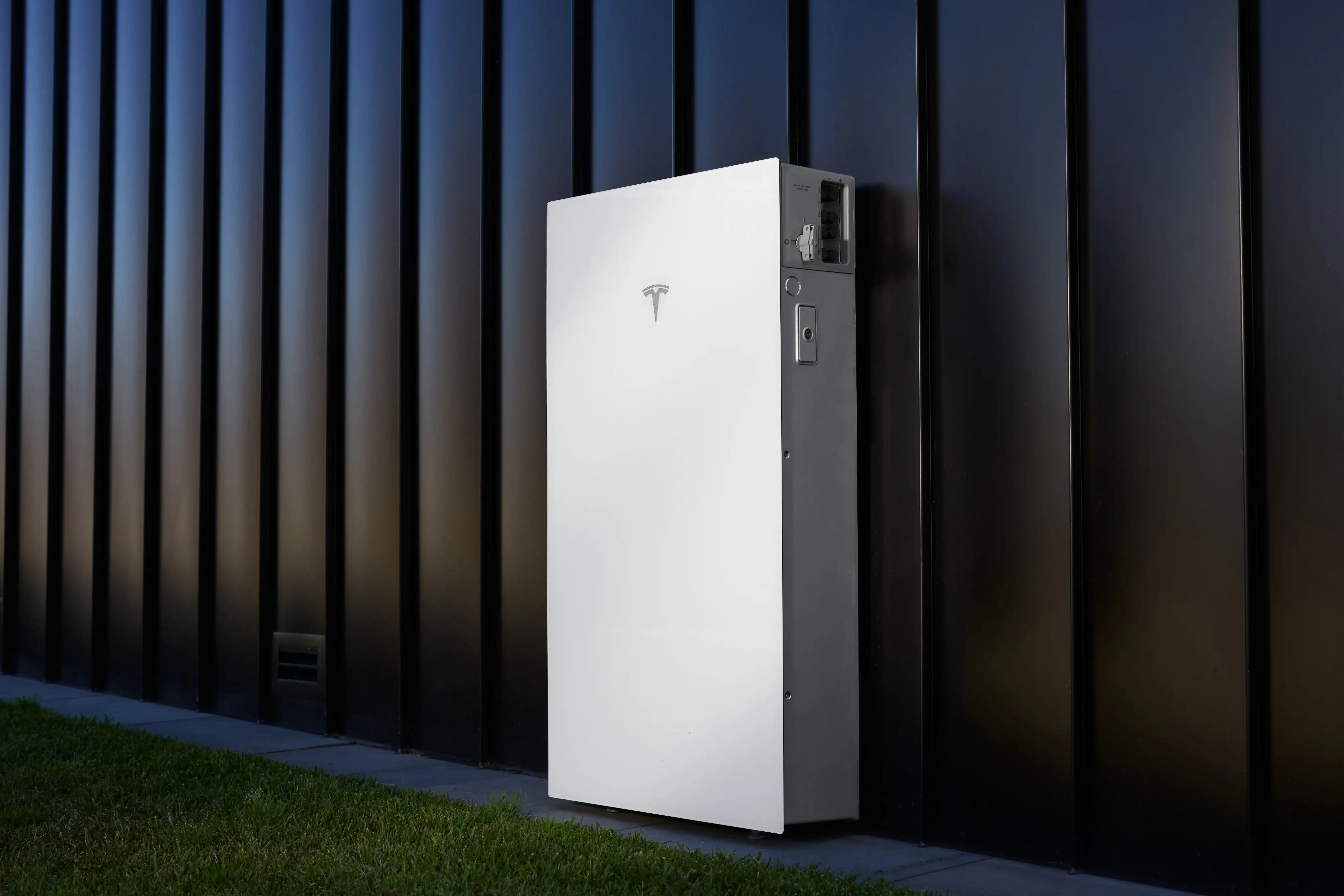 A white Tesla Powerwall 3P mounted on black vertical wall cladding beside a lawn