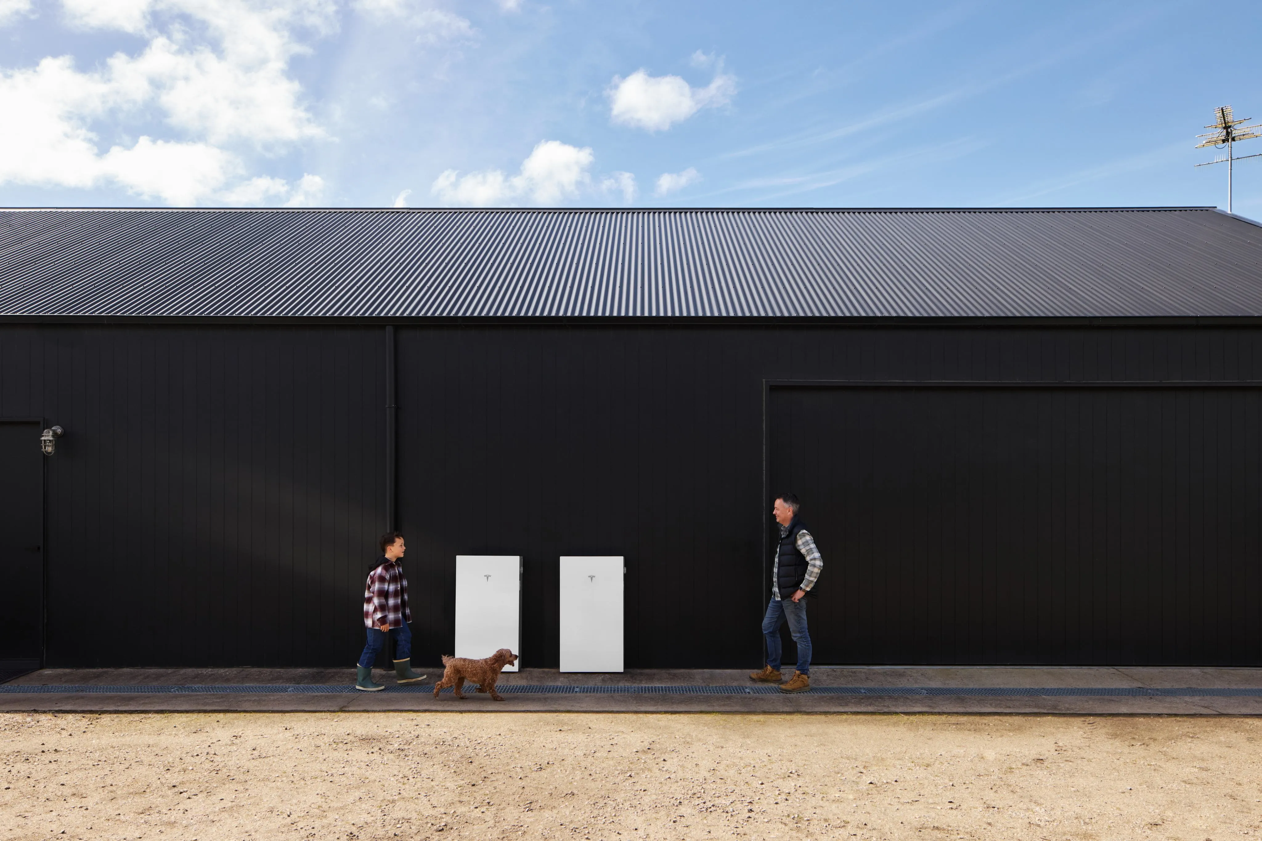 Two white Tesla Powerwall 3P units on the black wall of a long shed, a child with a dog on one side and a man walking on the other