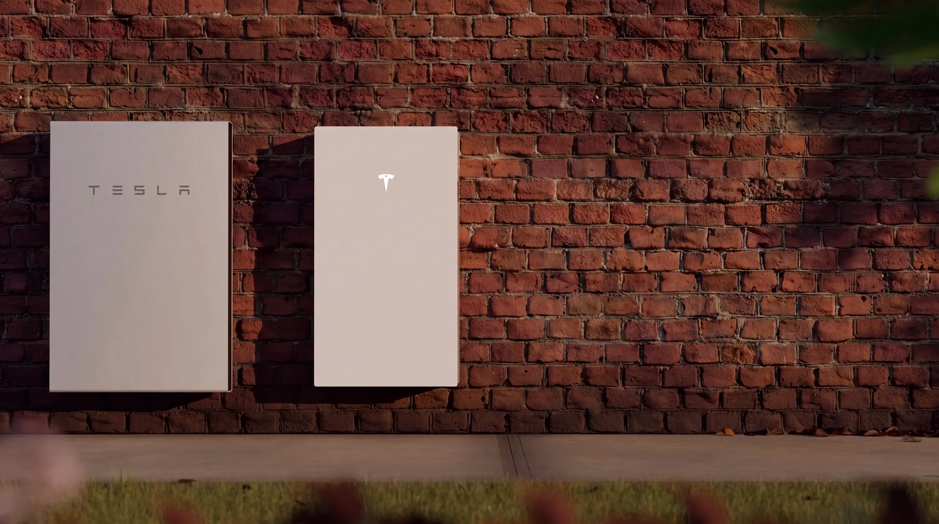 A Tesla Powerwall 2 and a Powerwall 3 mounted side by side on a red brick wall in afternoon light, the wider Powerwall 2 on the left and the narrower Powerwall 3 on the right