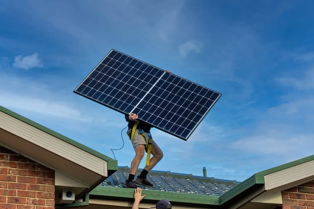 A person standing on a tiled roof holding a large solar panel above their head against a blue sky, with another person's hand reaching up from the gutter below