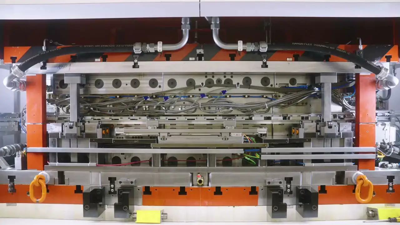 A close-up of press tooling on an automated production line, orange steel frames and hydraulic hoses over a row of machined plates