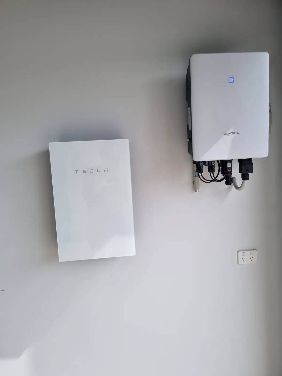 A Tesla battery and a Sungrow inverter mounted side by side on a plain garage wall.