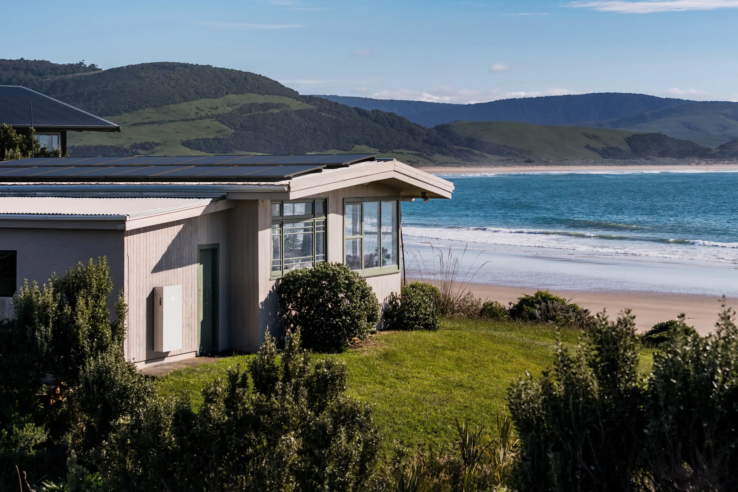 A weatherboard beach house with a full array of black solar panels on its low roof and a white home battery on the outside wall beside a green door, looking over grass to a beach and a headland