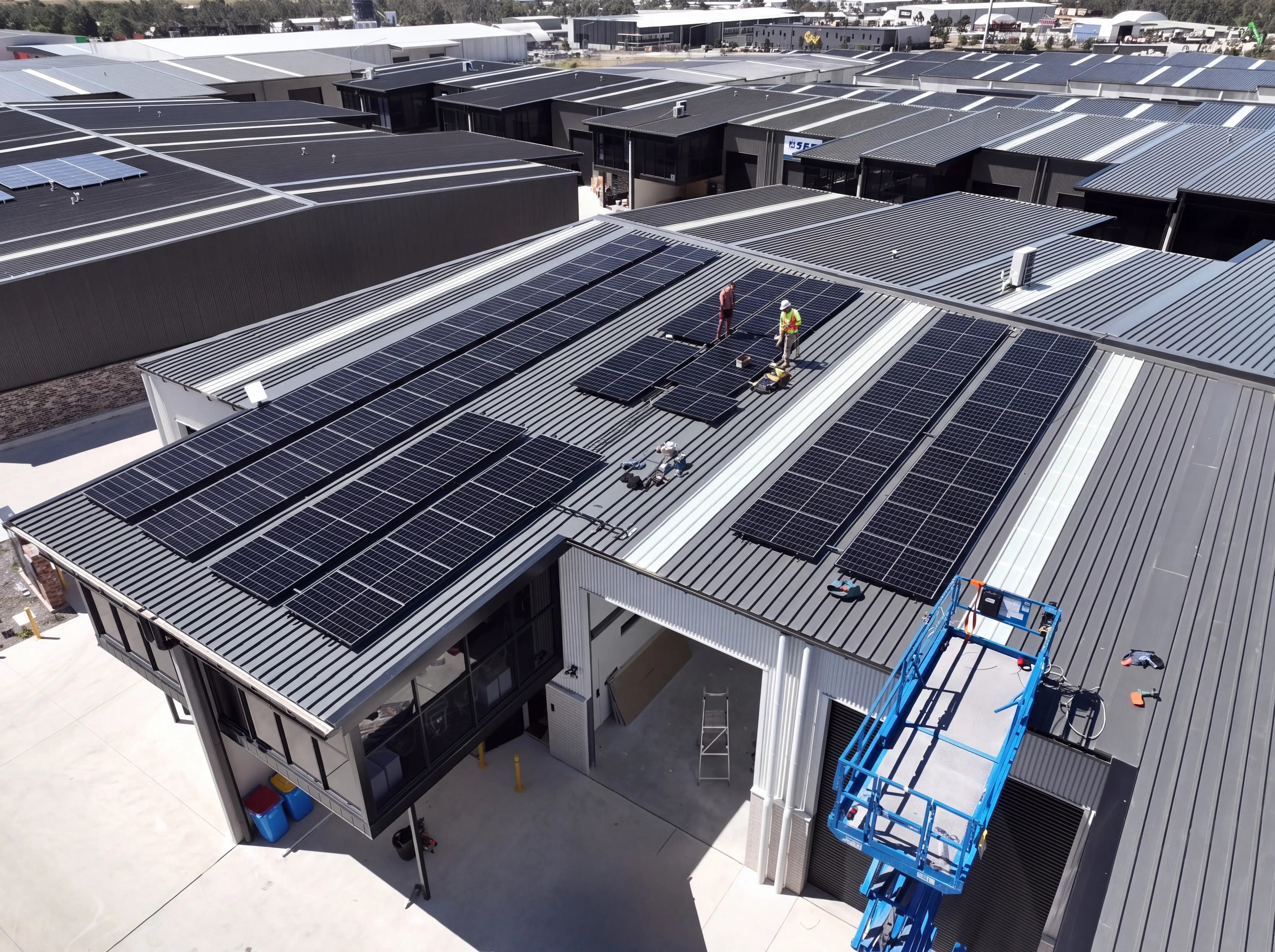 An aerial view of an industrial building with a dark ribbed steel roof carrying long rows of black solar panels, two workers in high-visibility clothing kneeling beside a part-laid section, and a blue elevated work platform at the roof edge
