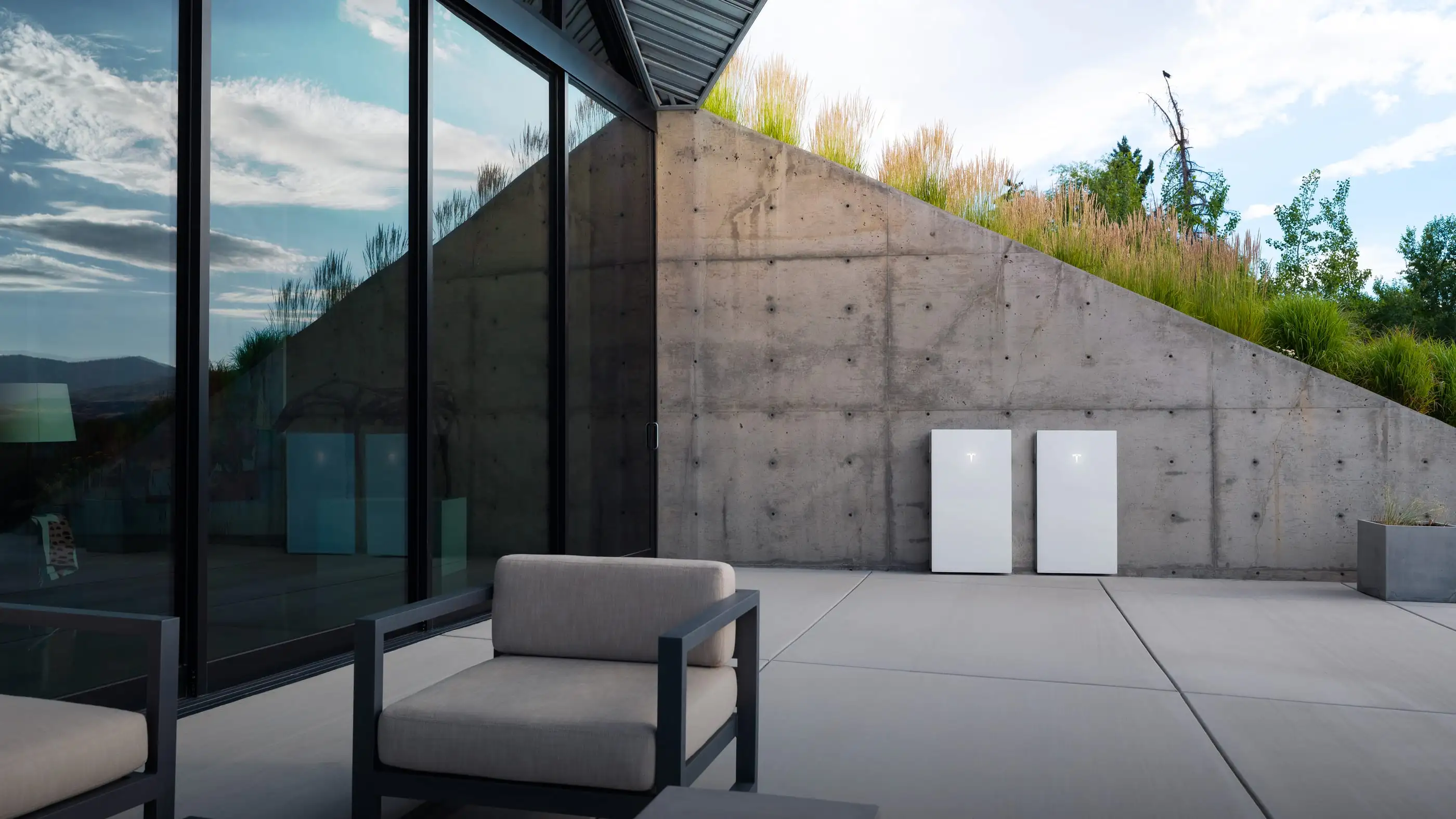 Two white Tesla home batteries standing side by side against a raw concrete retaining wall in bright daylight, with ornamental grasses along the top of the wall, the glazed corner of a house at the left and two upholstered chairs on the paving in front