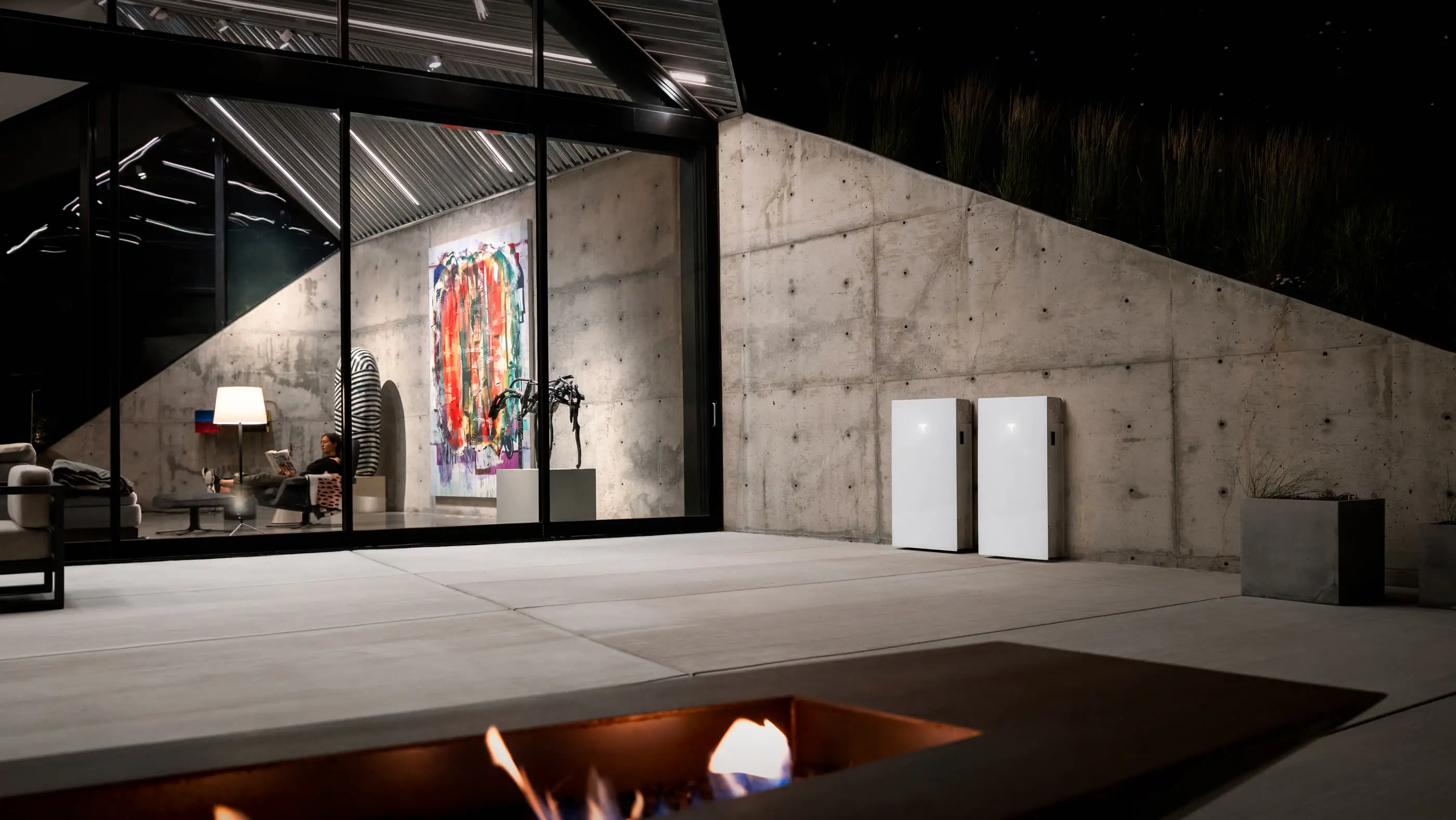 Two white Tesla home batteries standing against a raw concrete garden wall at night, beside the lit glass wall of a house with a painting and armchairs visible inside, and a gas fire trough burning in the paving in the foreground