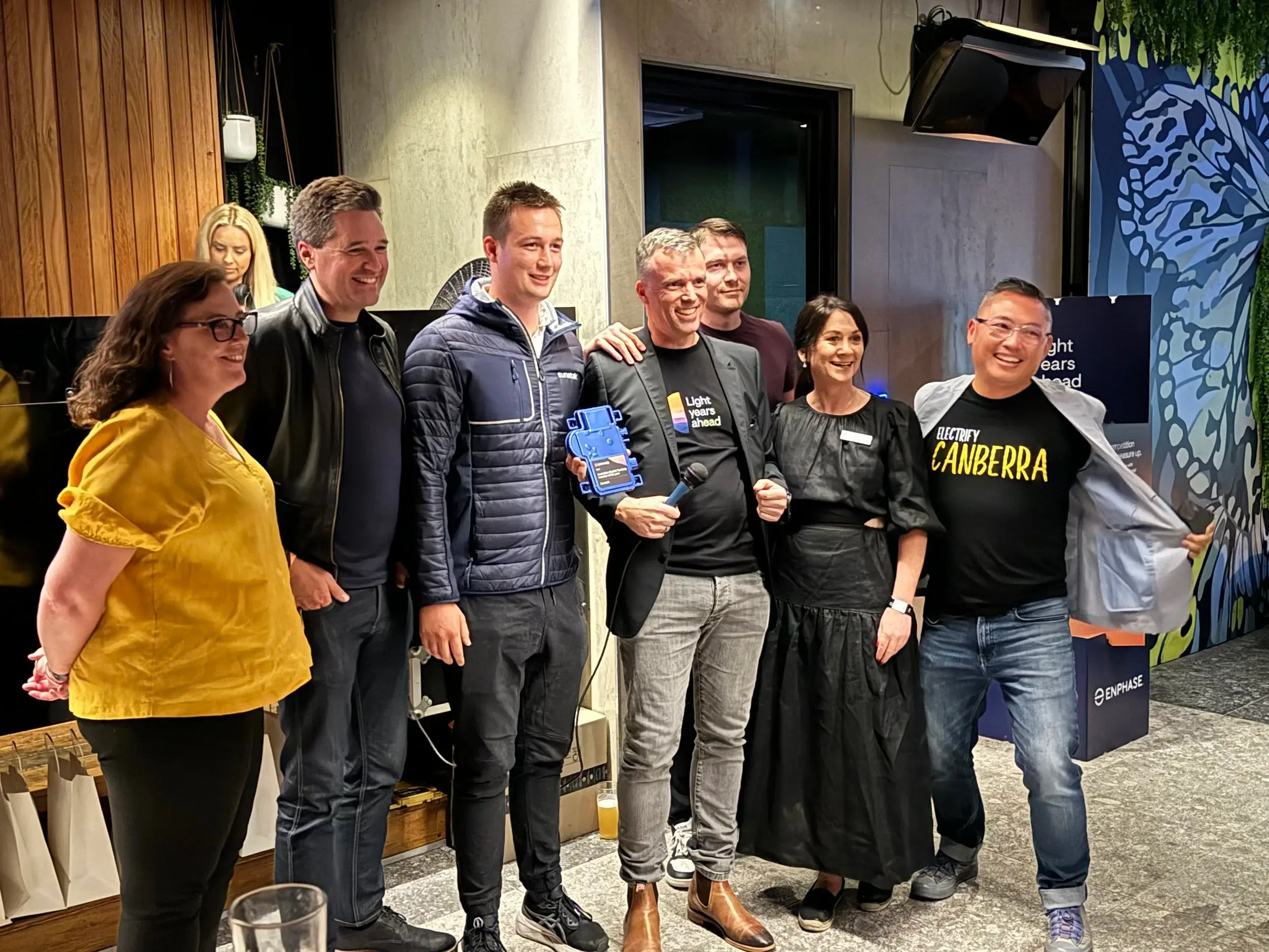 A group of Sunstak staff and guests at an indoor event, one holding a blue trophy and a microphone, with an Enphase-branded box and a shirt printed ‘Electrify Canberra’ among the group