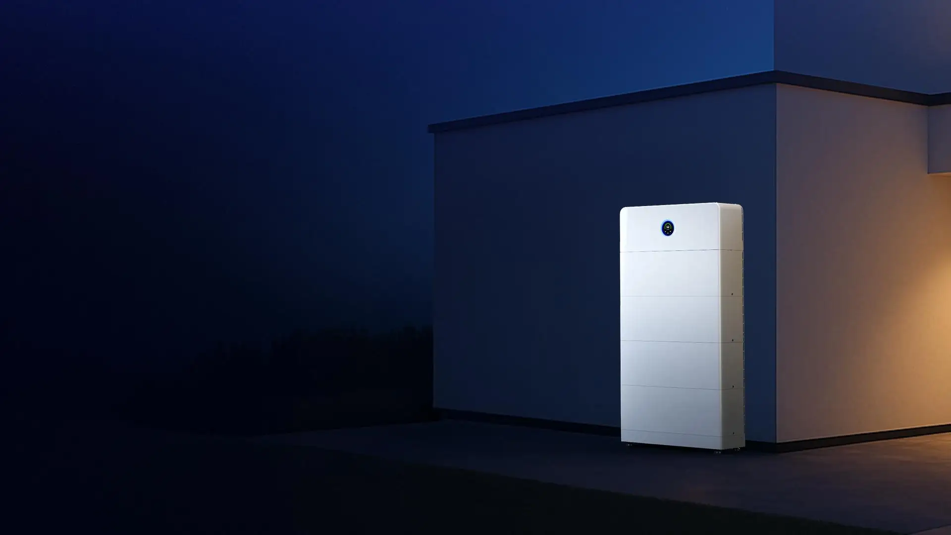 A white GoodWe battery, an inverter head over five battery modules, standing against the wall of a flat-roofed house at dusk with warm light falling on the wall beside it