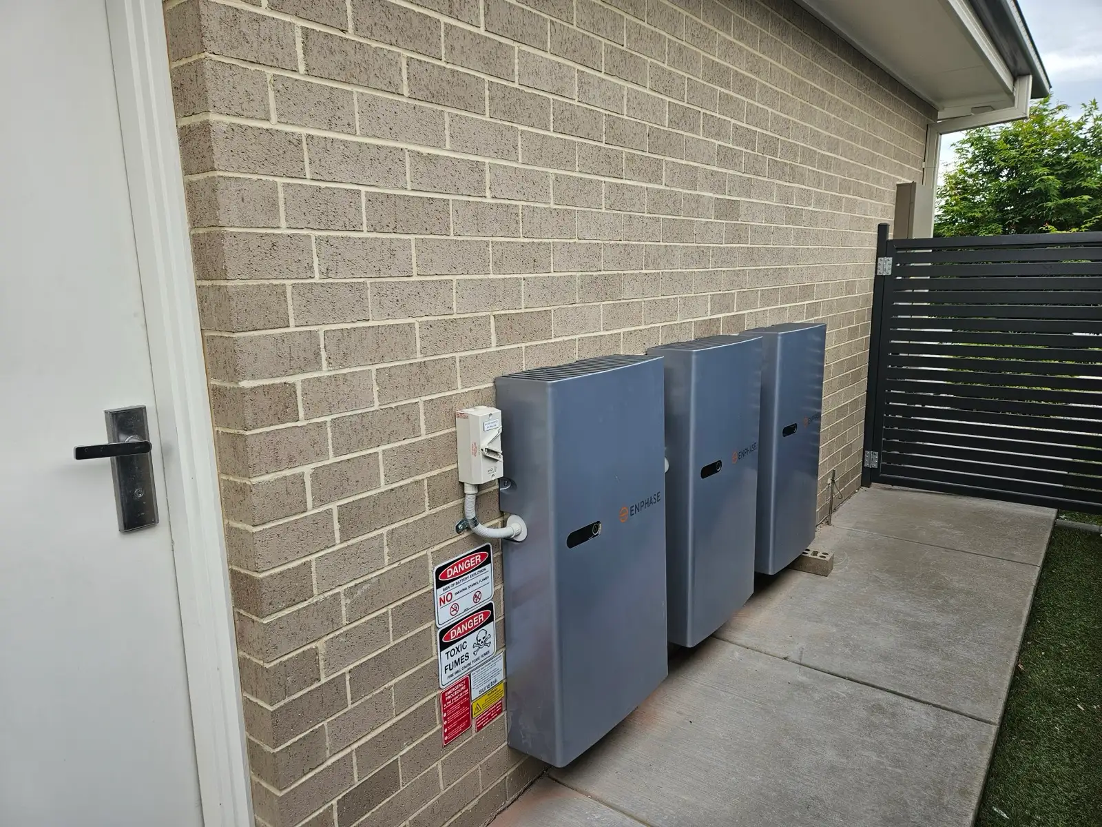 Three Enphase battery units lined up along a brick wall on a concrete path.