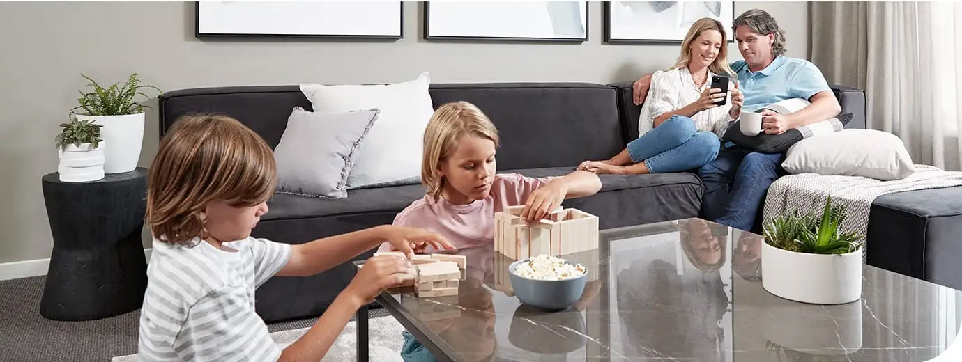 A family living room with two children building a block tower at a marble coffee table and two adults sitting together on a dark grey lounge behind them, framed prints on the wall and no air conditioning unit visible anywhere