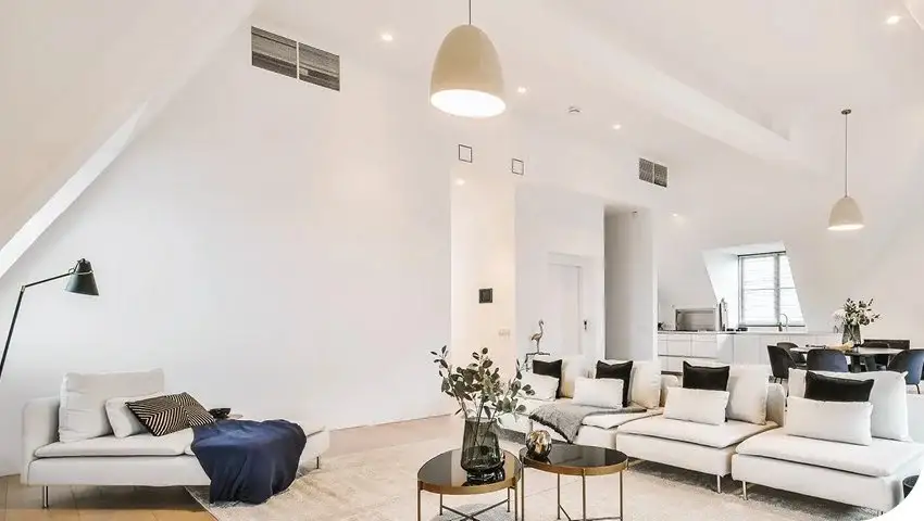 A white living area with a high sloped ceiling carrying two rectangular air conditioning vents, a pale sectional lounge below and a kitchen and dining table visible beyond