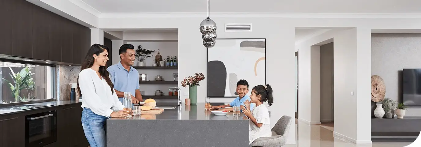 An open-plan kitchen and living area with a rectangular white air conditioning vent set into the ceiling above the island bench, two adults standing at the bench and two children sitting at it, and no unit visible on any wall
