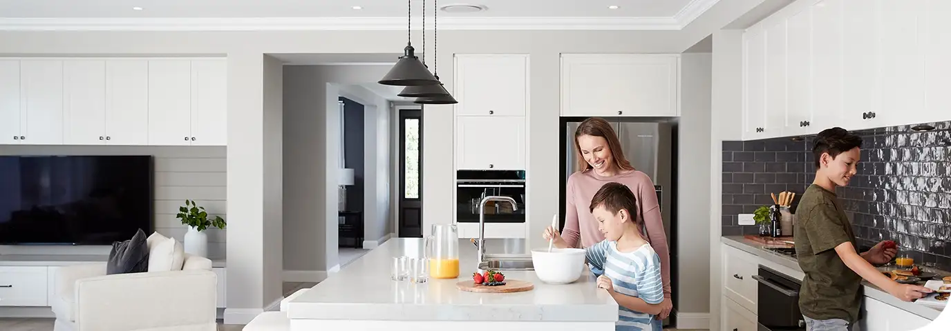 A wide open-plan kitchen and living area with a white island bench, a woman and a boy mixing something in a bowl at the bench, a second child at the counter beyond, and a lounge and television at the far left