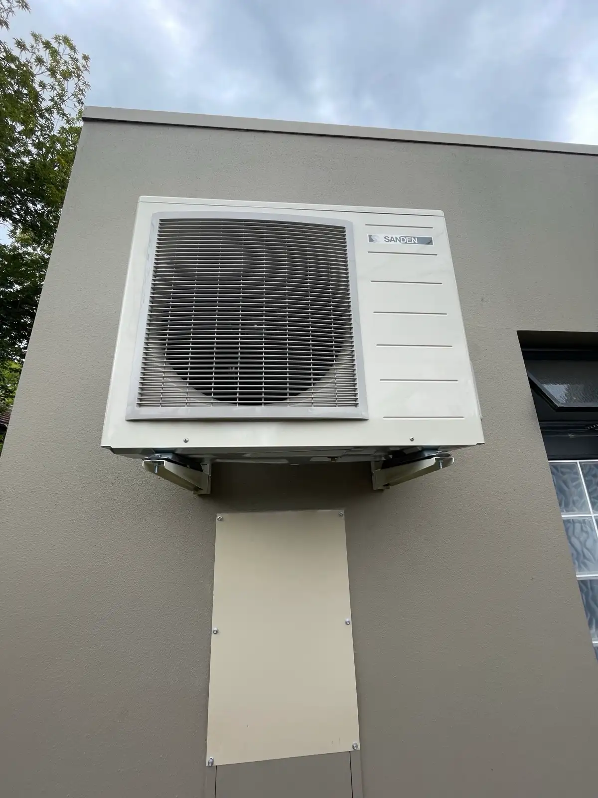 A Sanden heat pump unit bracketed to a rendered wall, its pipework boxed in beneath.