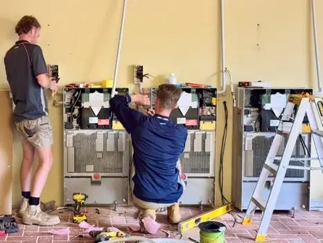 Two people working on three wall-mounted units with their covers off, showing the wiring inside, with a stepladder and tools on the floor