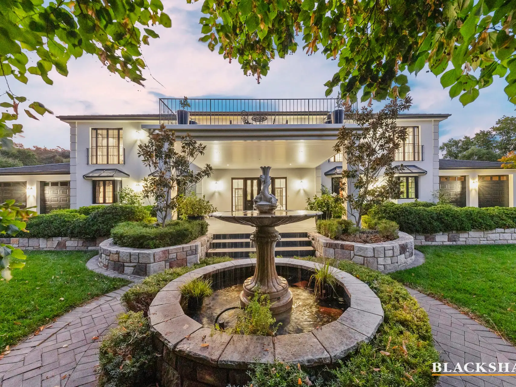 The front of a large white two-storey house at dusk, lit from inside and out, with a stone fountain in a circular pond on the lawn in front of it, a flight of steps up to a columned entrance, clipped hedges either side and a tree in leaf overhanging the frame
