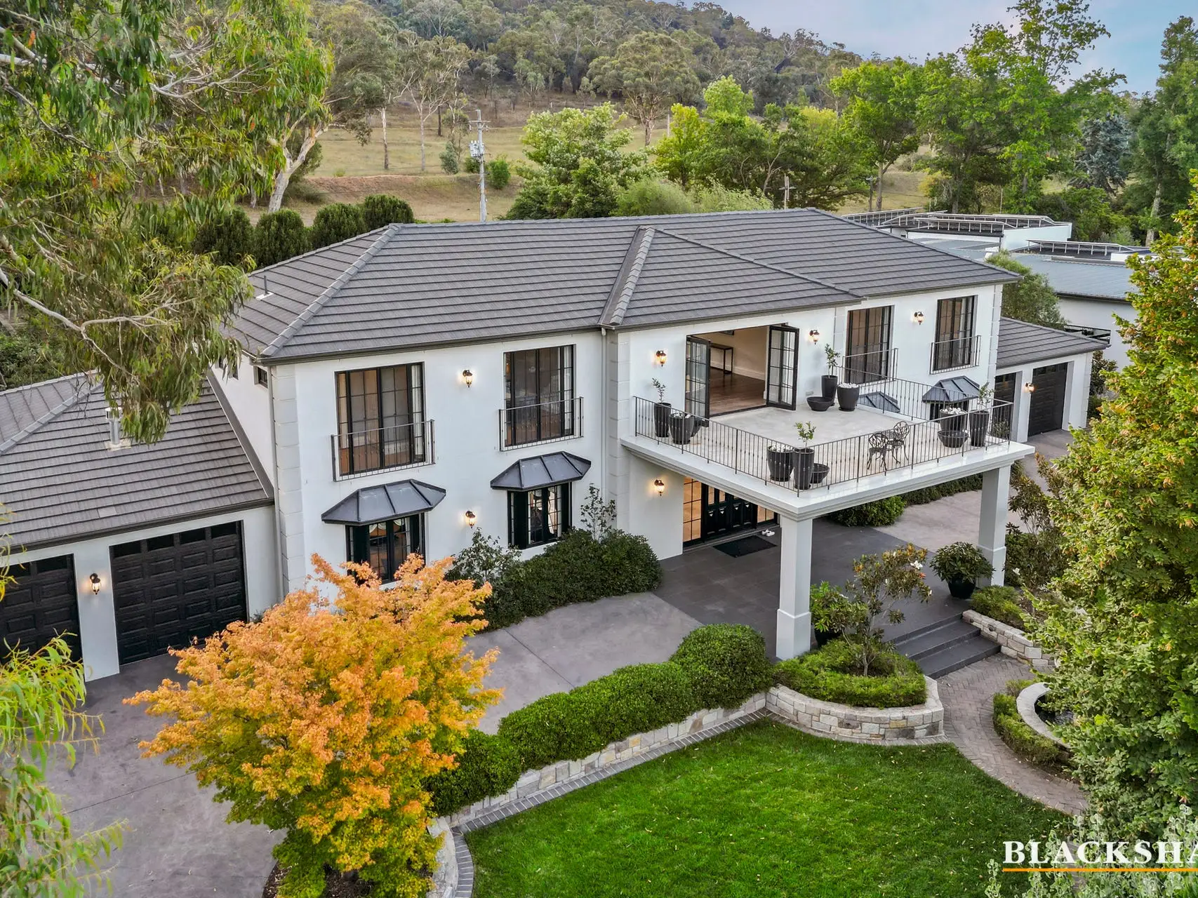 A rendered two-storey house photographed from above the front garden, with a balcony across the upper floor, two dark garage doors at ground level, clipped hedges and a lawn in front of it, a maple in autumn colour beside the drive and open hillside behind