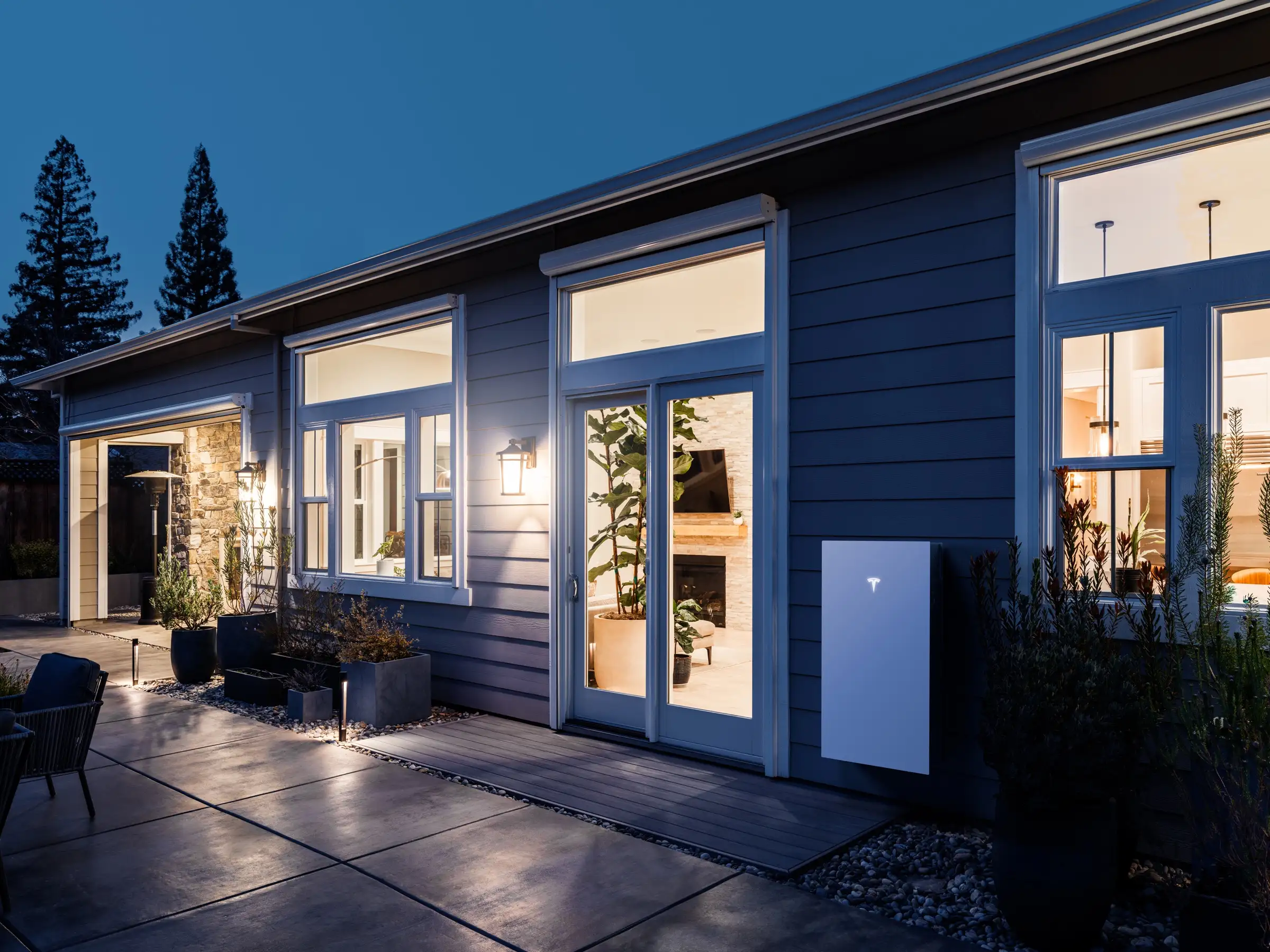 The side of a single-storey house at dusk, lit from inside through large windows, with a white home battery mounted on the dark blue cladding beside a glazed door
