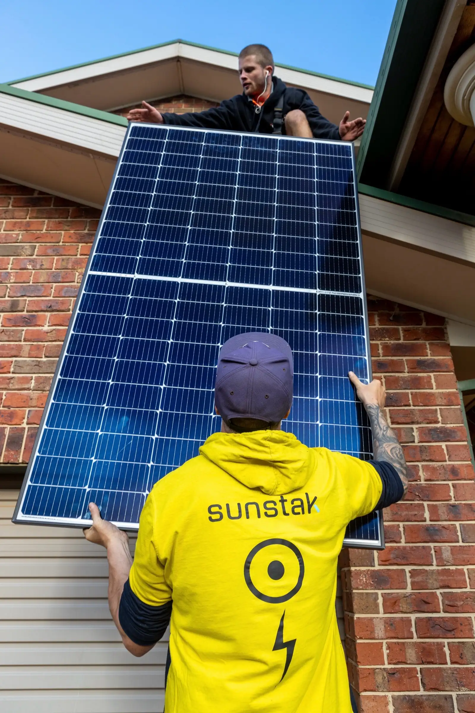 Seen from below, a person in a yellow sunstak hoodie and a navy cap holds a large solar panel flat against a brick wall while a second person leans down from the roof edge above with both arms out to take it, against a blue sky
