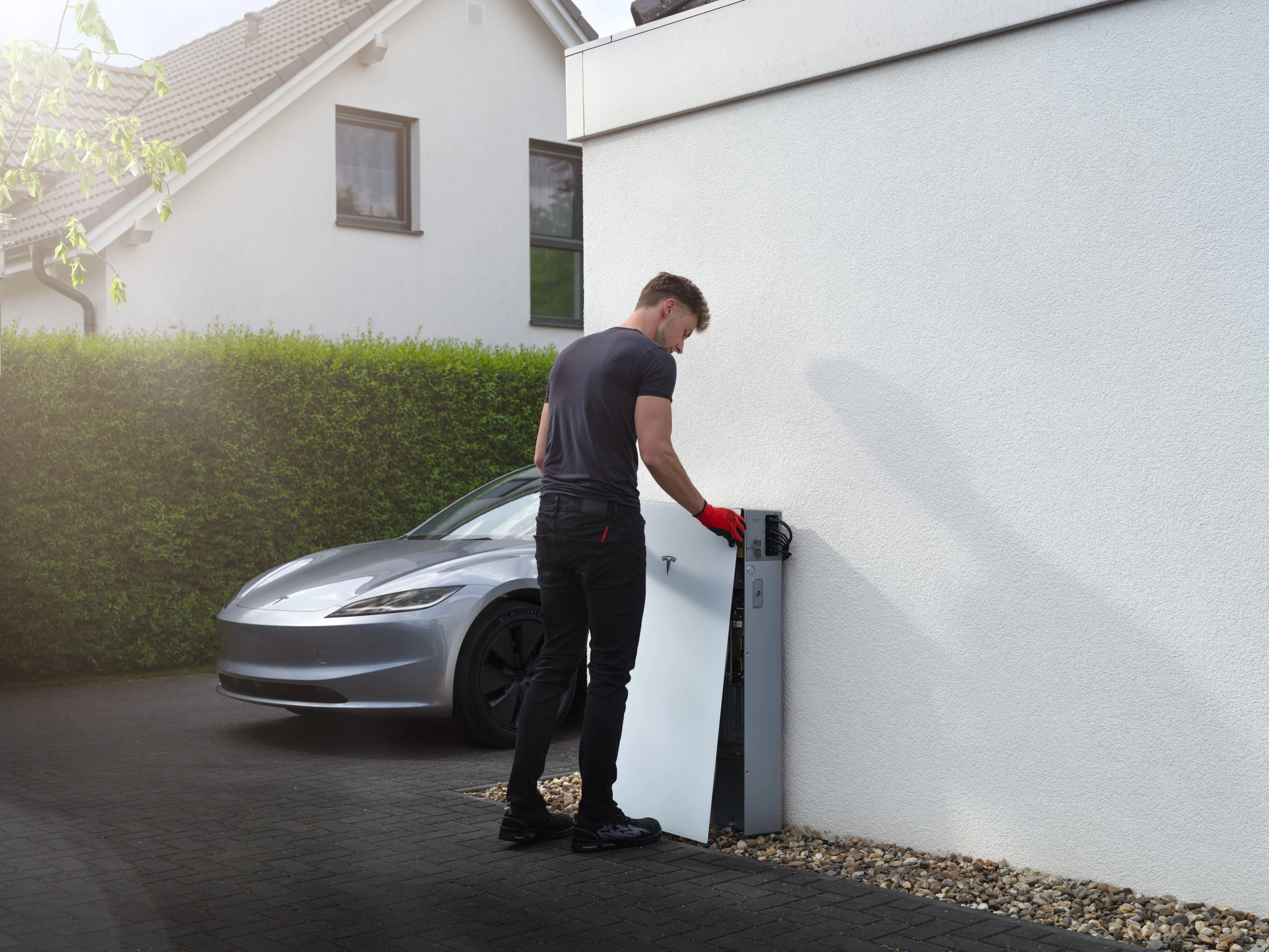 An installer in red gloves fitting a white Tesla Powerwall 3P to a white rendered wall, a silver car parked on the driveway behind him
