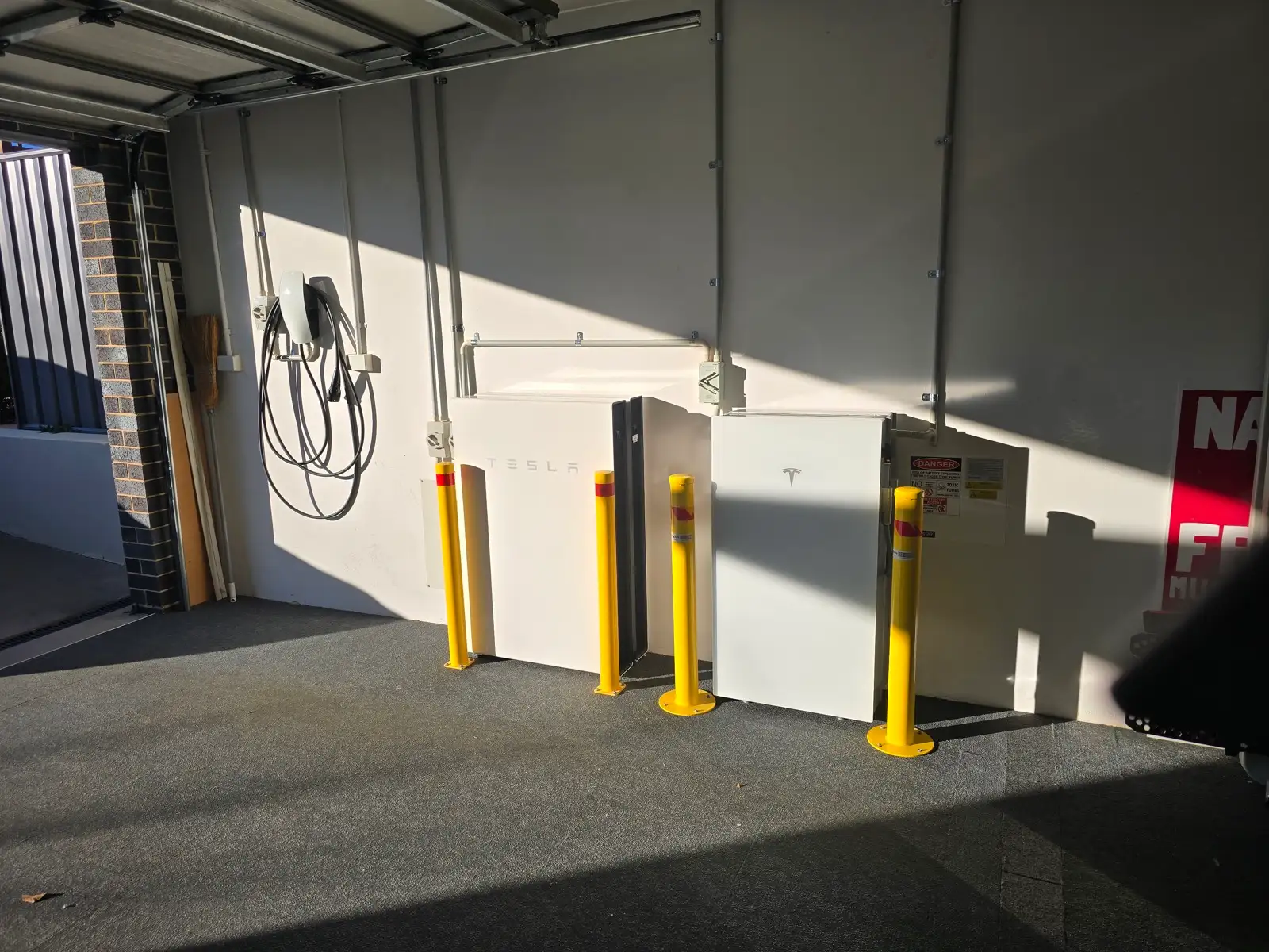 A garage bay with two Tesla batteries behind bollards and a Tesla wall connector coiled on the wall.