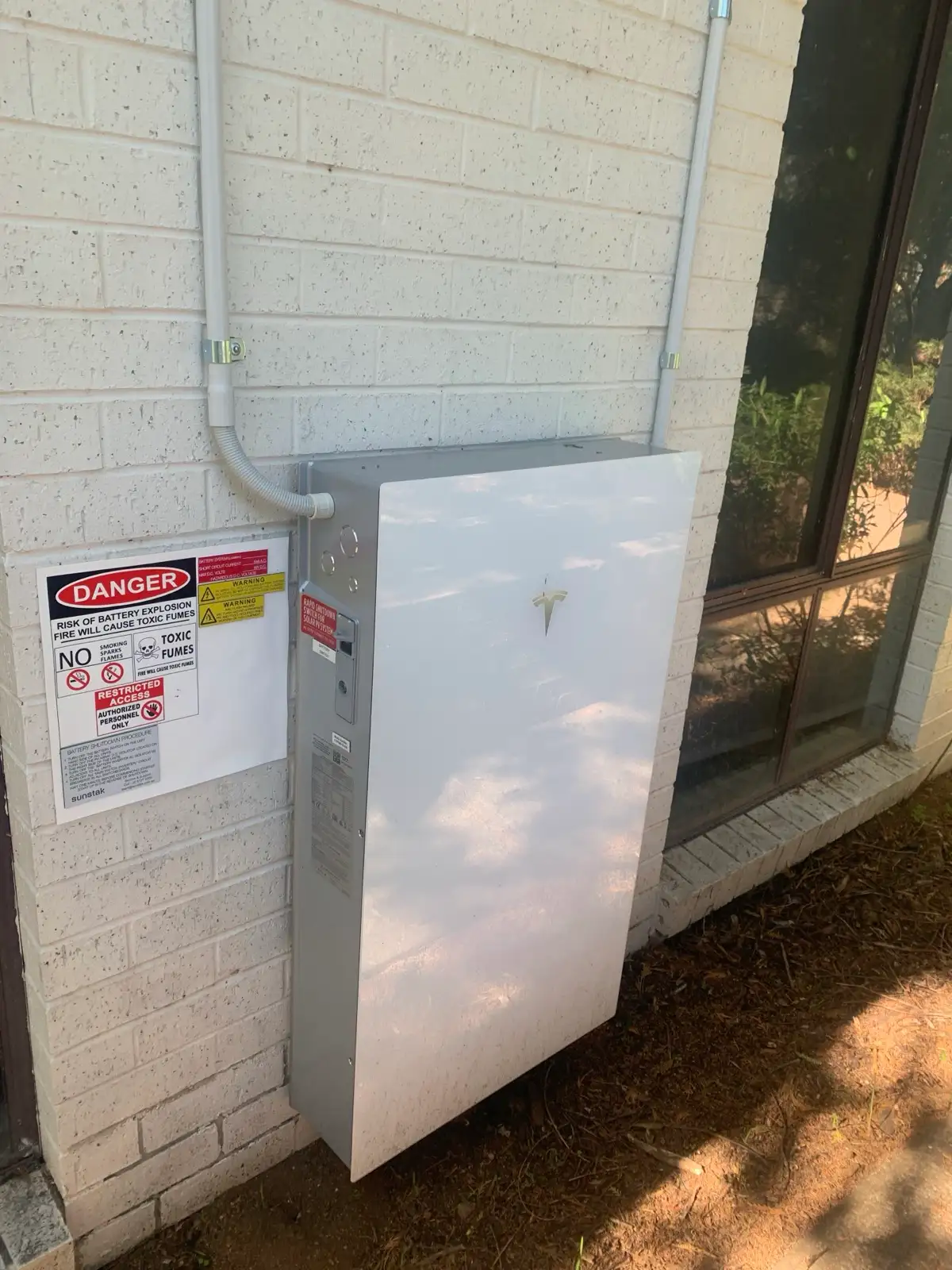 A Tesla battery standing against a painted brick wall beside its shutdown placard and conduit.