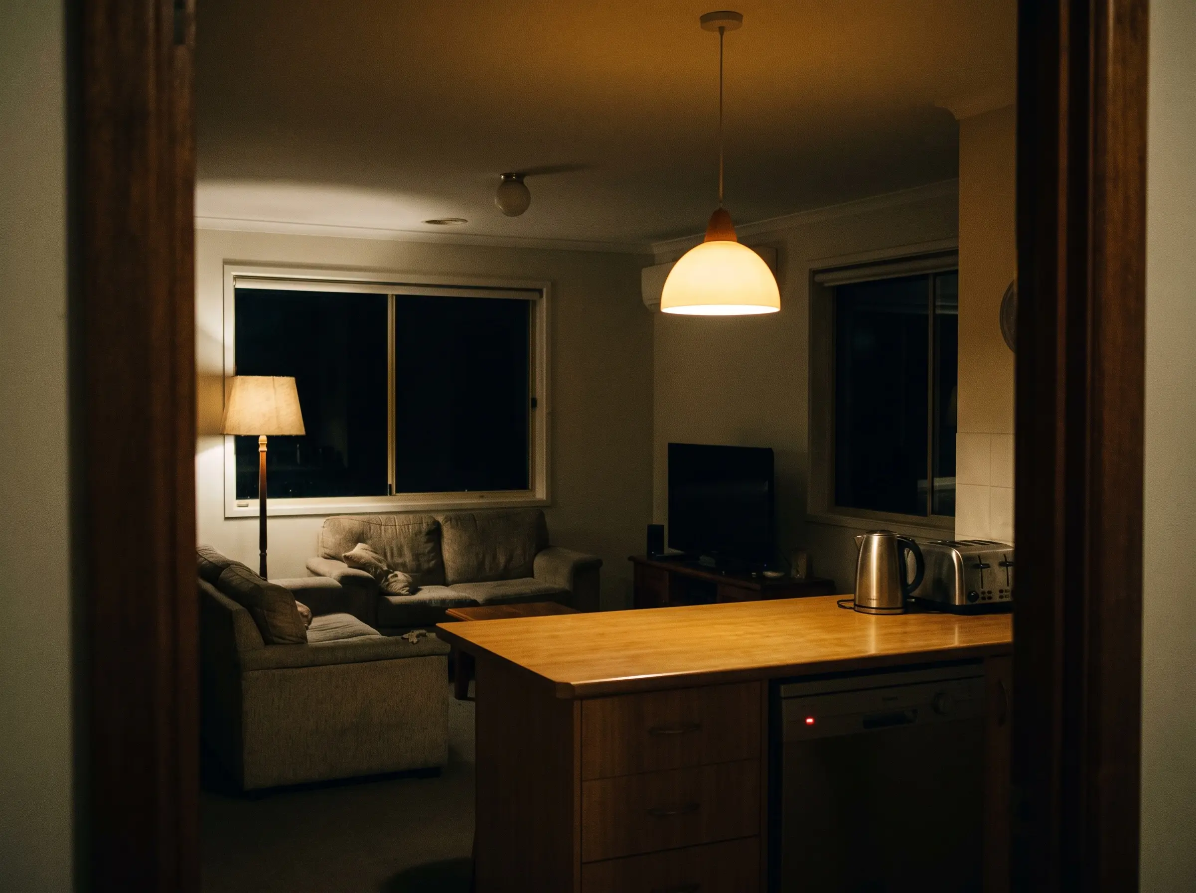 A kitchen bench and a living room at night with two lights on, a kettle and a toaster on the bench, a dishwasher indicator lit below it and a dark television beyond