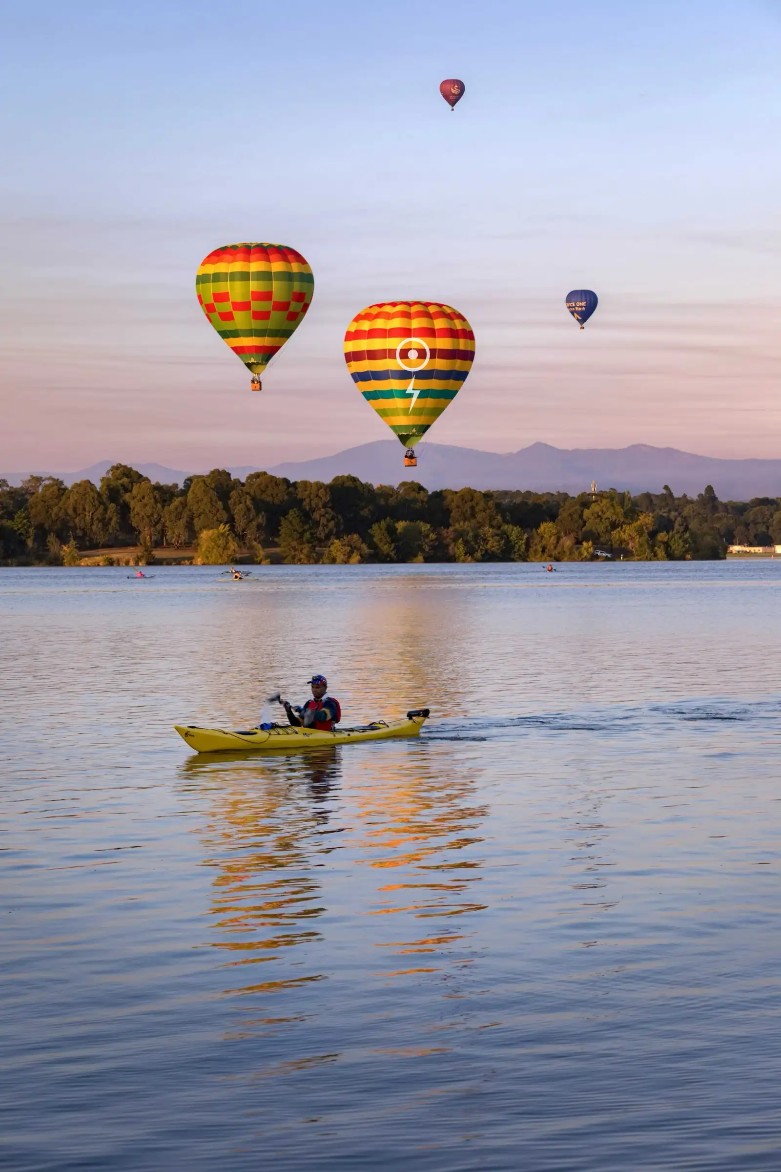 Hot-air balloons over still water at dawn, with a distant treeline and low hills behind them