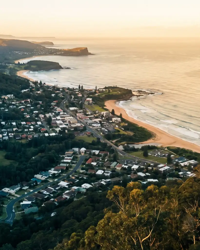 Sunset over a coastline seen from a forested escarpment: a small town on flat ground between the slope and the sea, a sand beach, and headlands stepping away into the distance