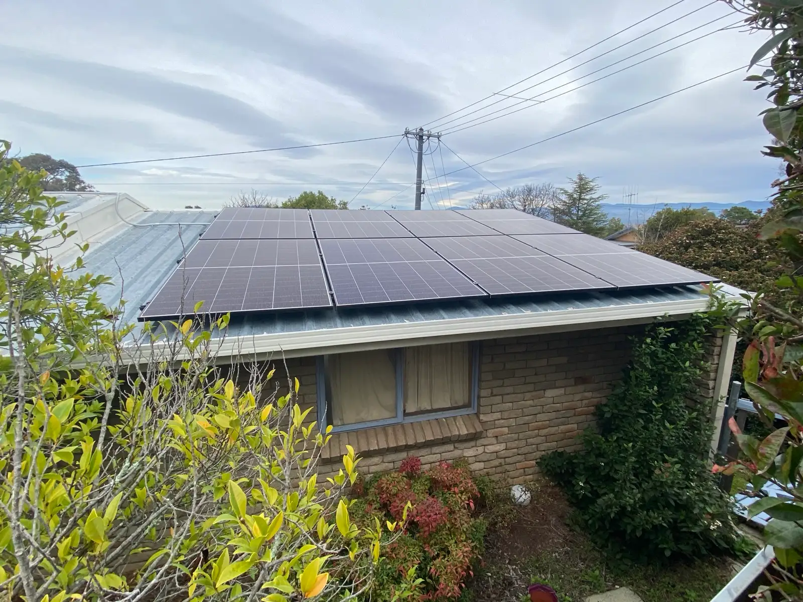 Solar panels squared off across the corrugated roof of a brick house, hills on the skyline behind.