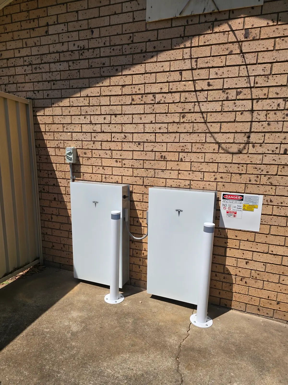 Two Tesla batteries on a concrete slab against a brown brick wall, a white bollard in front of each.