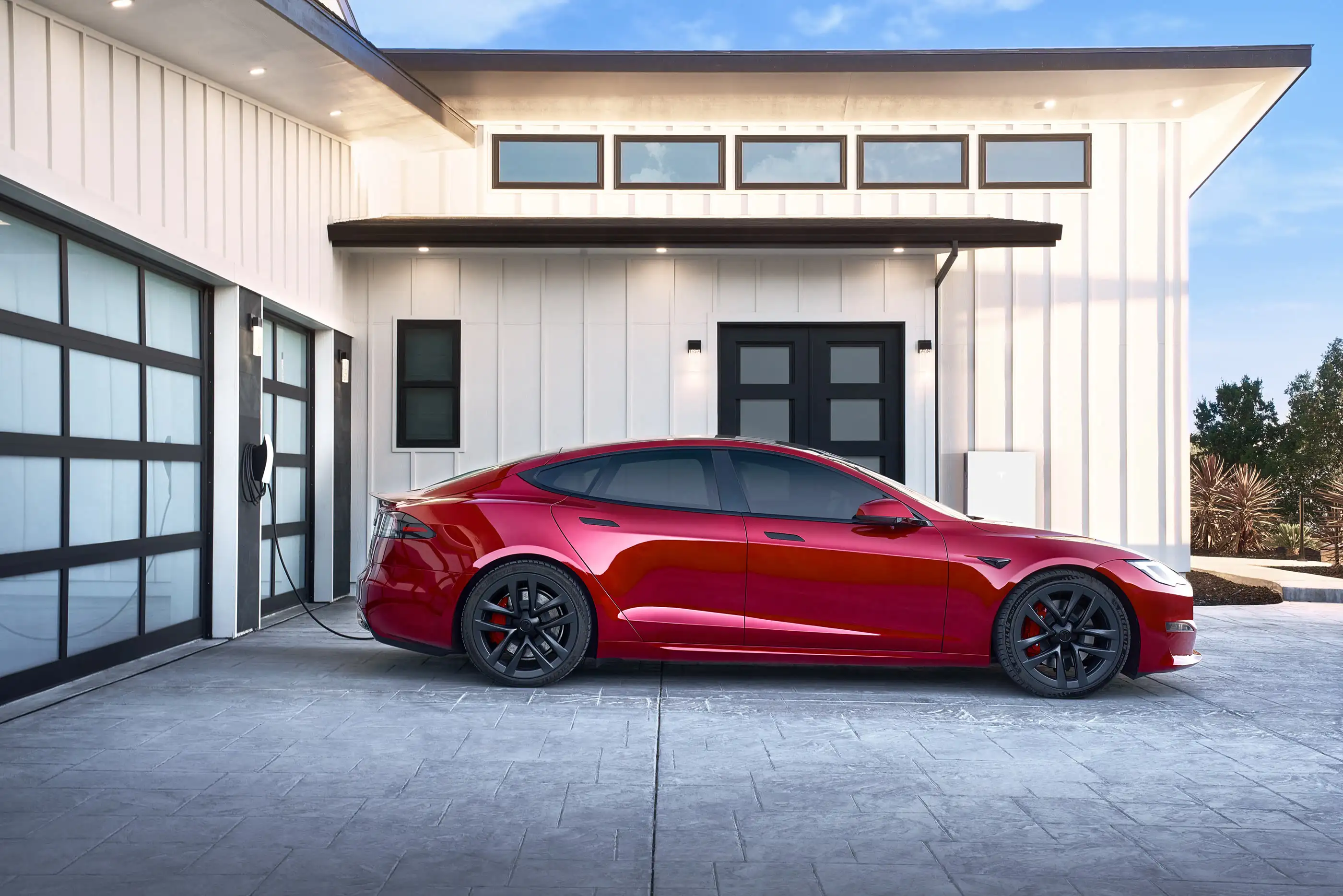 A red Tesla Model S parked on a paved driveway beside a white house, its charging cable running back to a Tesla Wall Connector on a dark pillar between two glass garage doors.