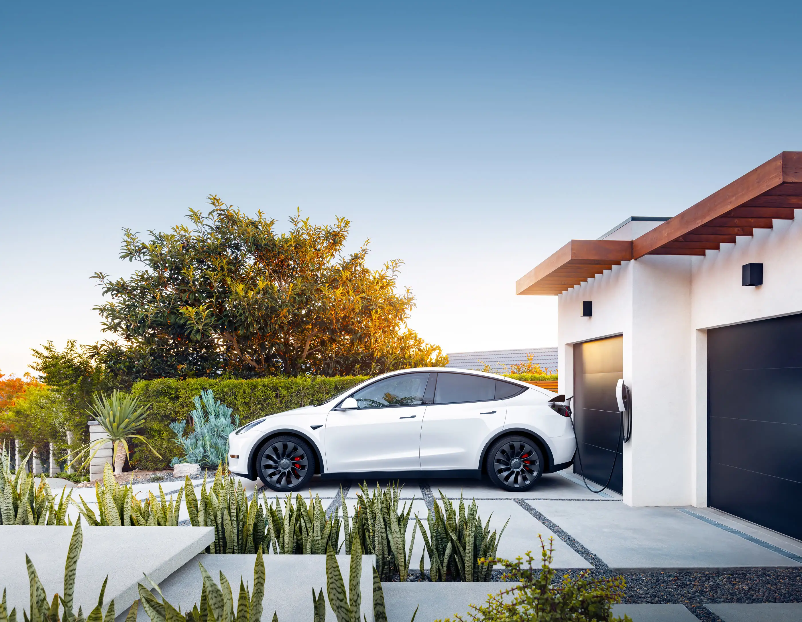 A white Tesla Model Y parked side-on outside a white rendered house at dusk, its cable running to a small white charger mounted on the wall between two dark garage doors, with agaves and a clipped hedge in the foreground