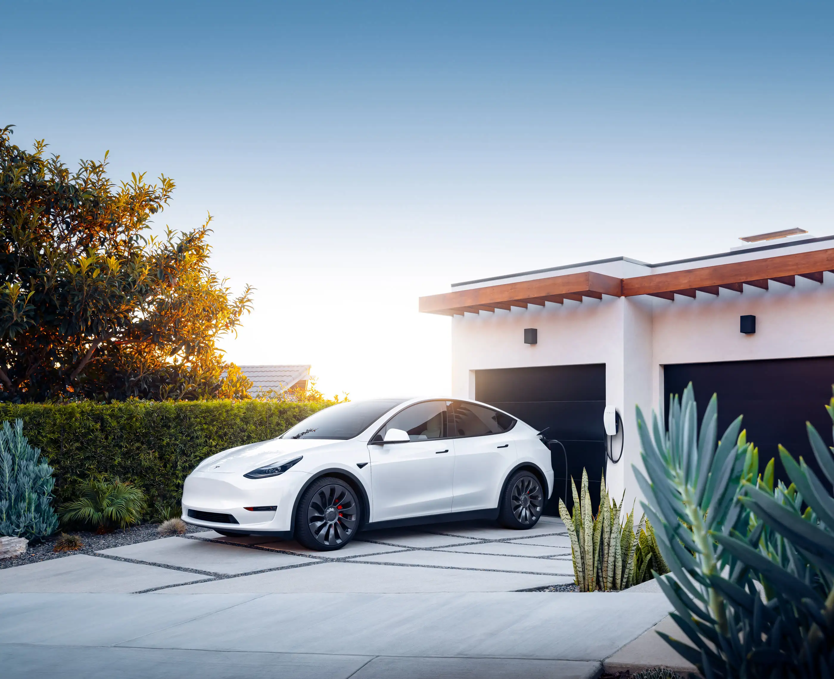 A white Tesla Model Y on a paved driveway at sunset, angled towards the camera in front of a white house with two dark garage doors and a timber-lined eave