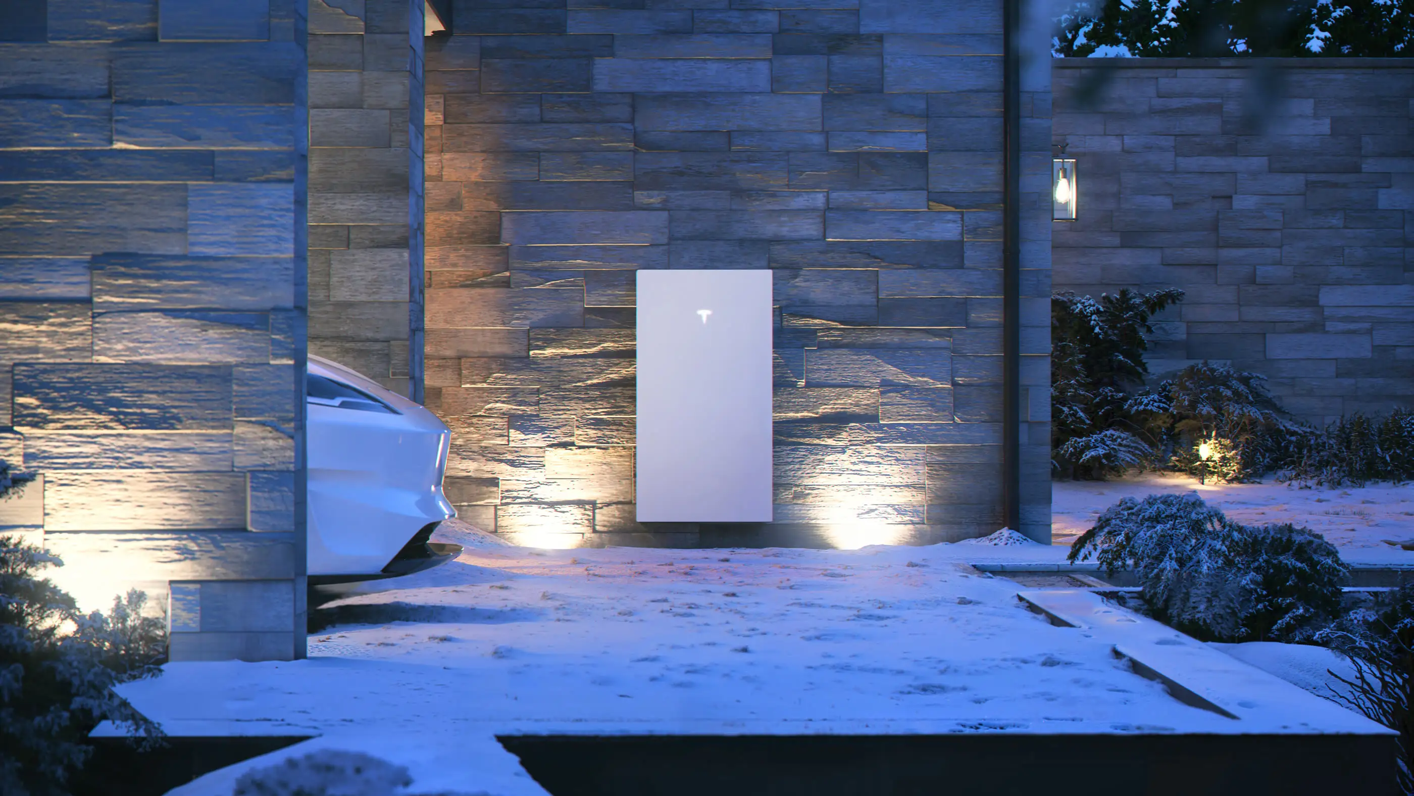 A white home battery on a stacked stone wall at night, uplit from below, with snow on the ground and on the shrubs beside it and the nose of a white car at the left
