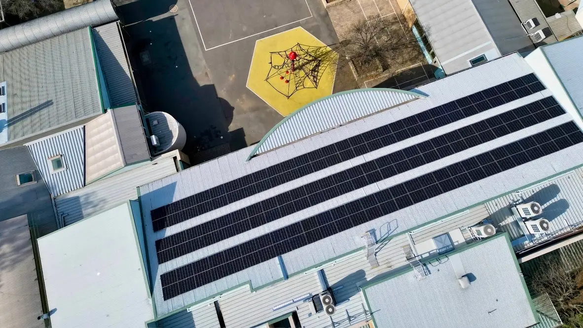 Overhead aerial view of four long rows of solar panels on the curved-ridge roof of a large school building, with a playground and a yellow climbing frame in the yard alongside