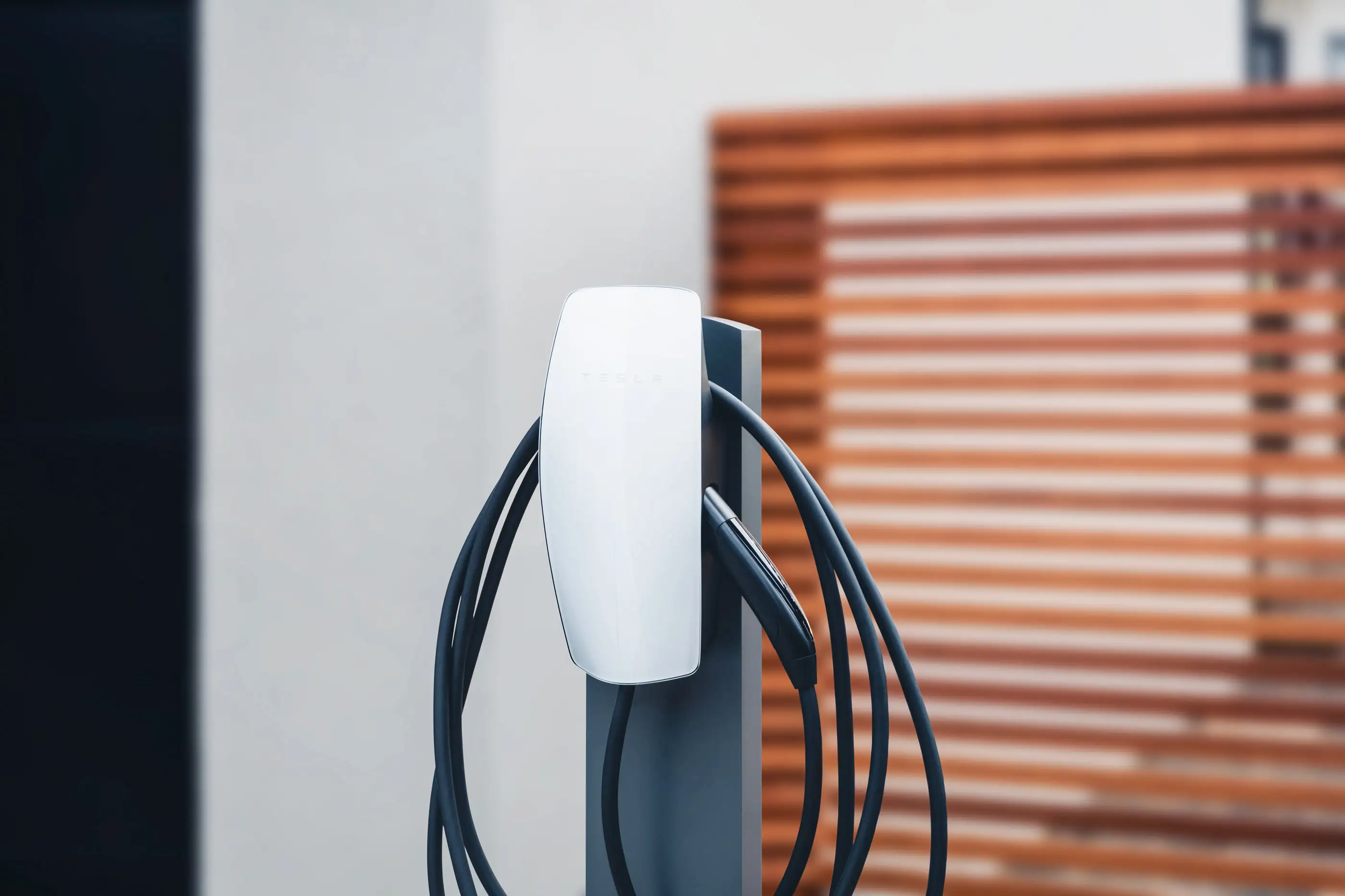A white Tesla Wall Connector on a grey steel pedestal with its cable coiled around it, standing on paving in front of an out-of-focus timber slat screen
