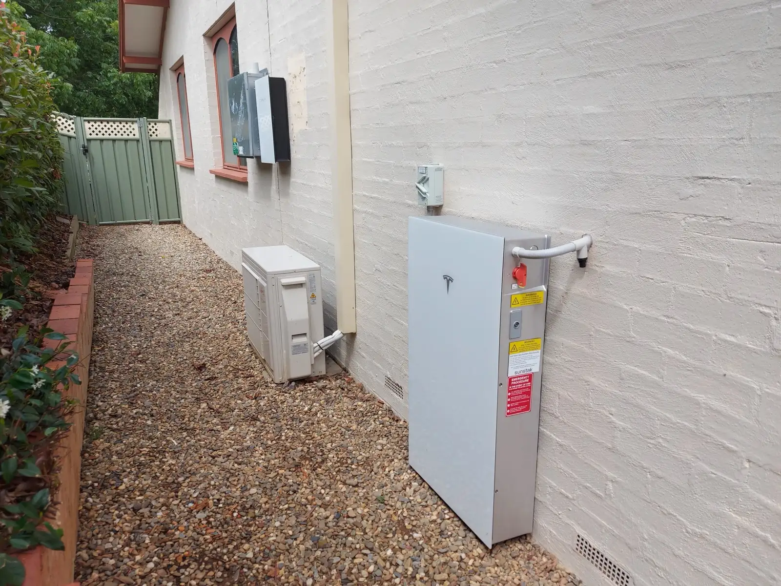A Tesla battery standing on gravel against a painted brick wall, the air conditioner and meter further along.