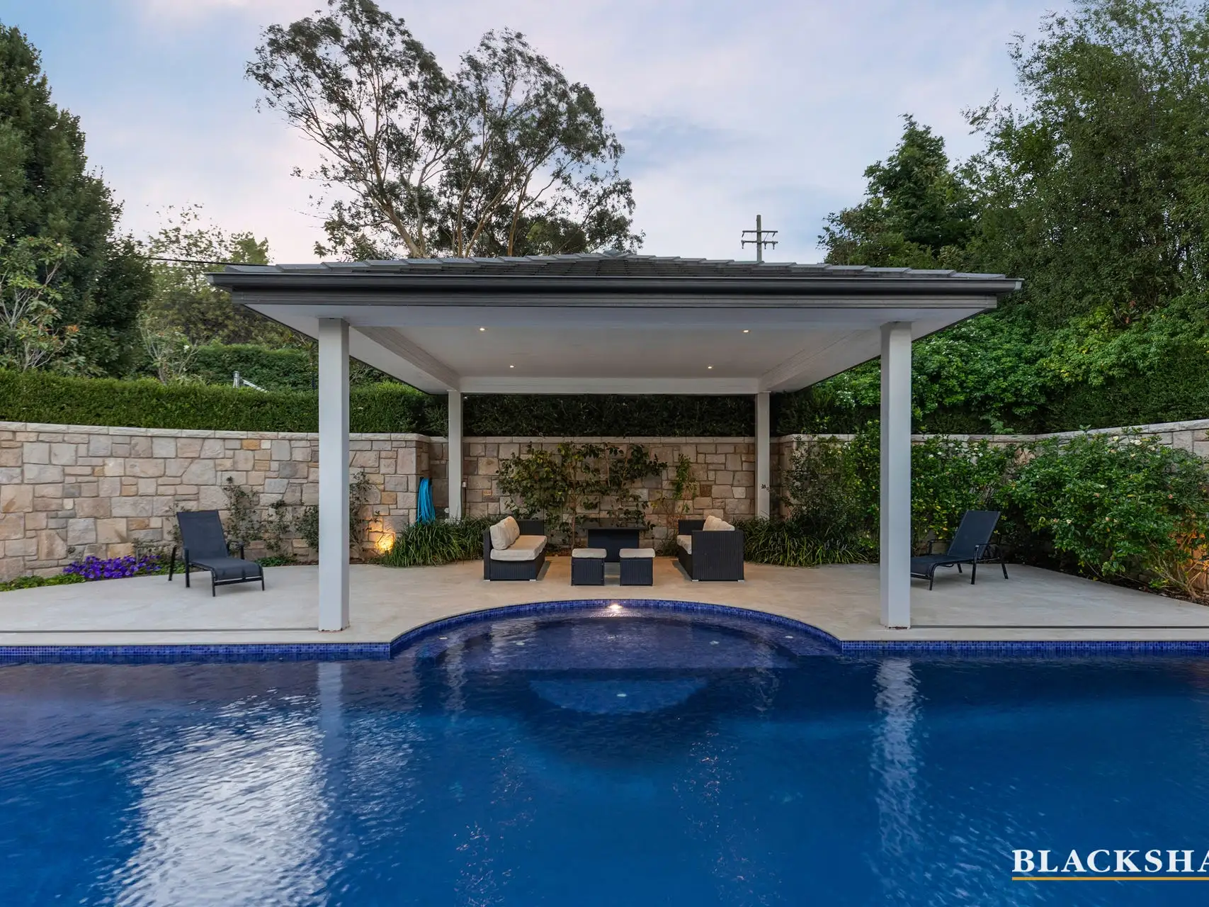 A swimming pool at dusk with a rounded end, an open-sided pavilion with a flat roof and lounge seating under it standing behind the water, a coursed sandstone wall and clipped hedge along the boundary and a gum tree against a pink sky