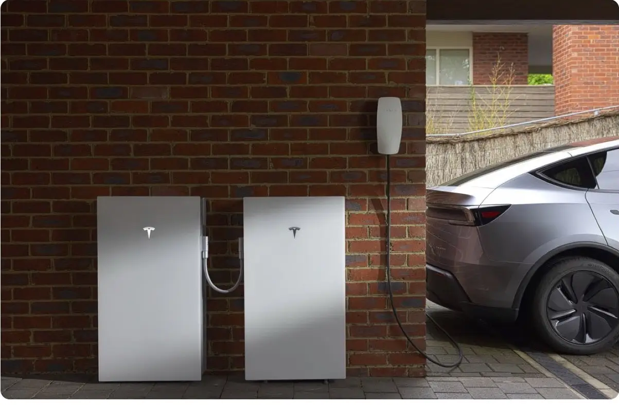 A Tesla battery mounted on a brick wall beside a car charging on a driveway.