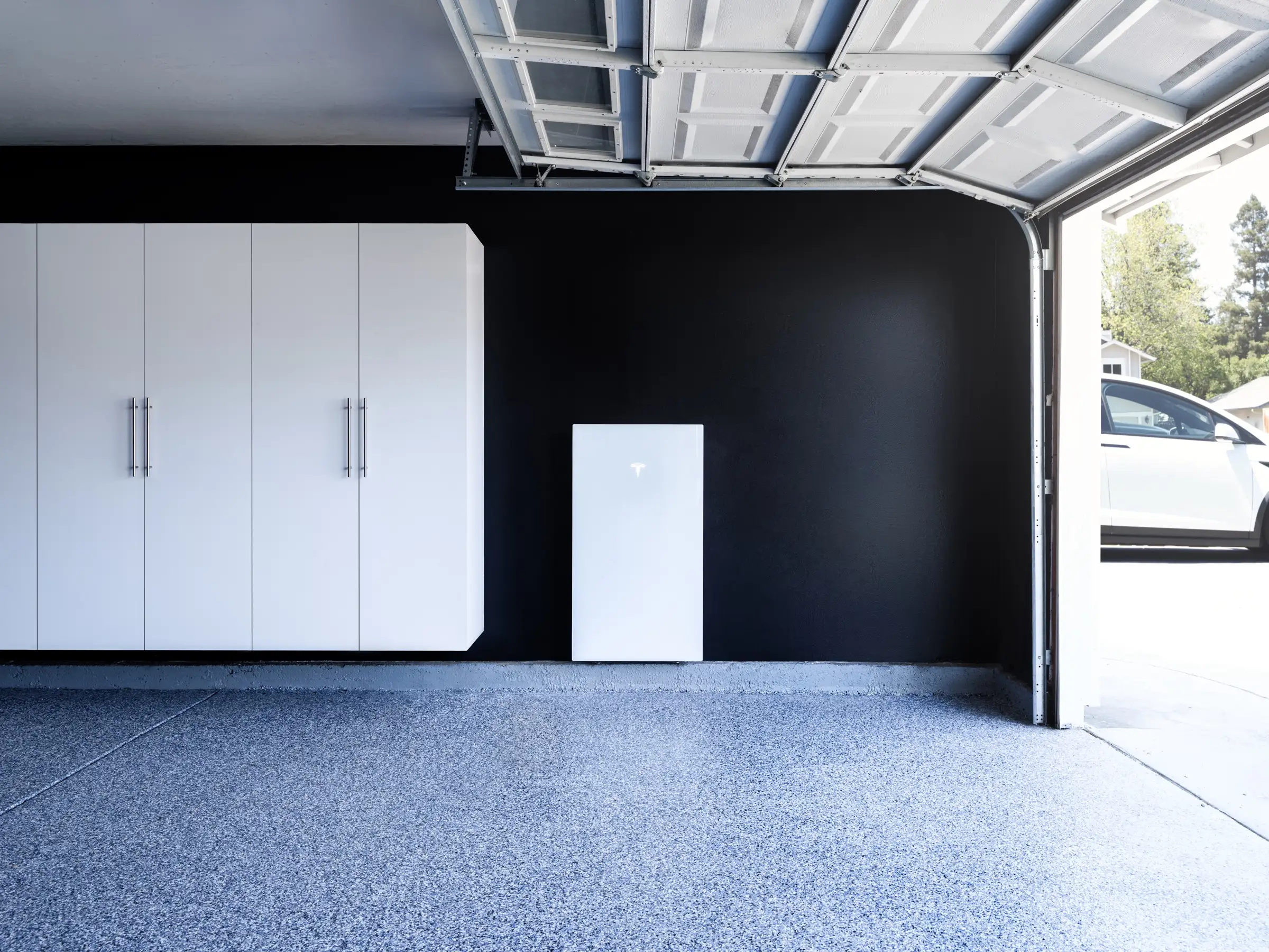 A white home battery standing against the black-painted back wall of an open garage, with white cupboards to the left, a speckled concrete floor and a white car on the driveway outside