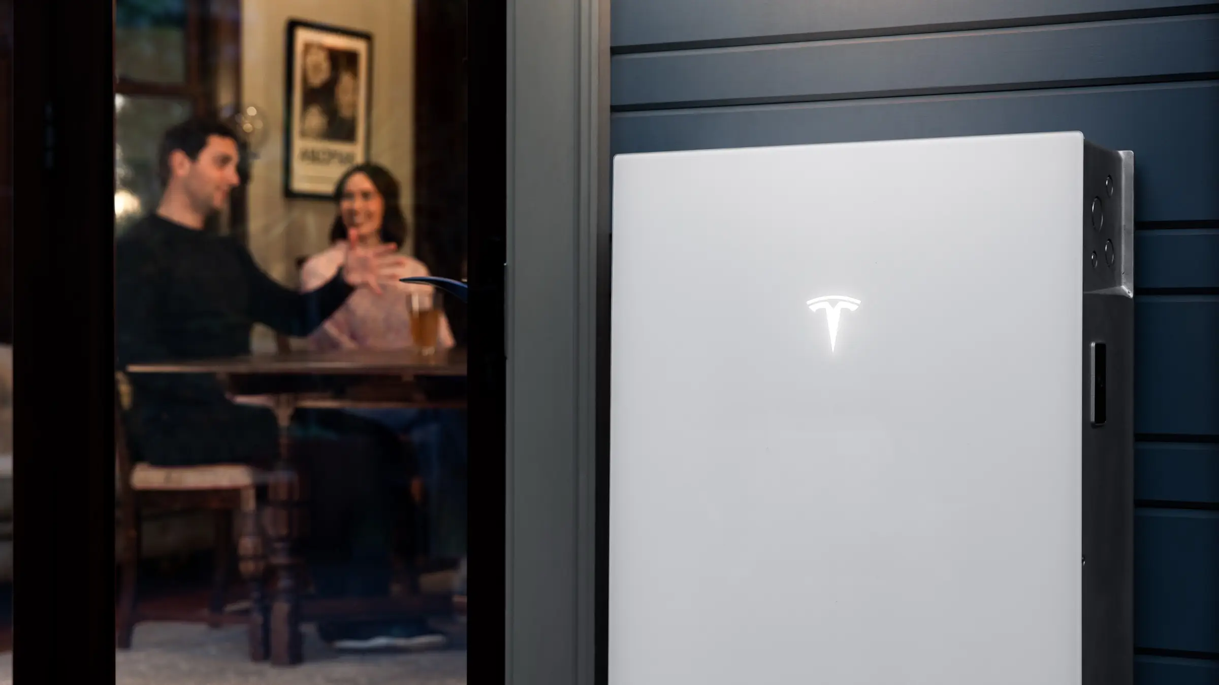 A white Tesla home battery mounted on dark blue weatherboard beside an open door, with two people talking at a table inside the lit room