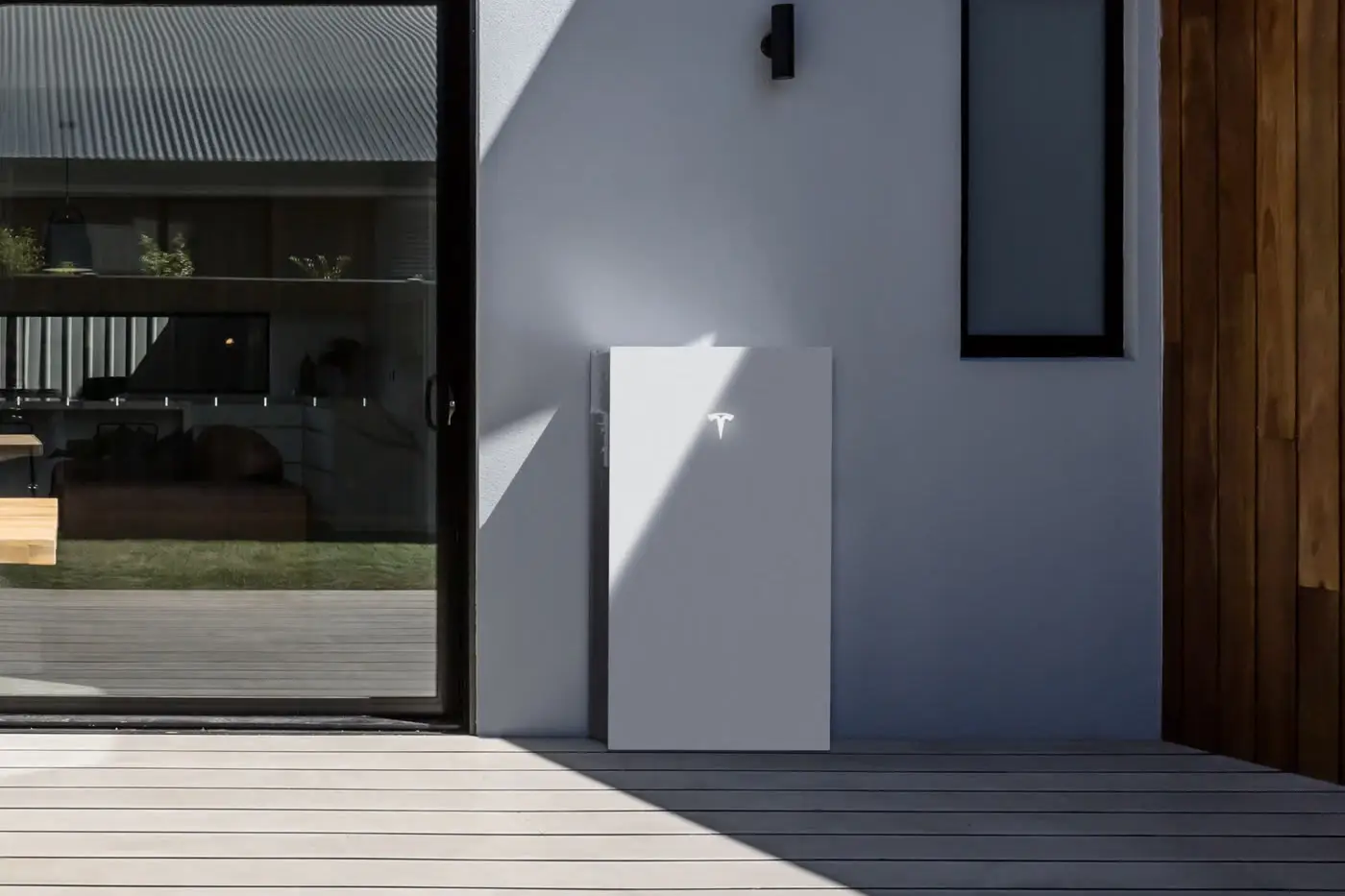 A single white Tesla-badged battery mounted on the grey rendered wall of a house, beside a timber-clad corner and a tall narrow window, standing on a pale timber deck in hard sunlight
